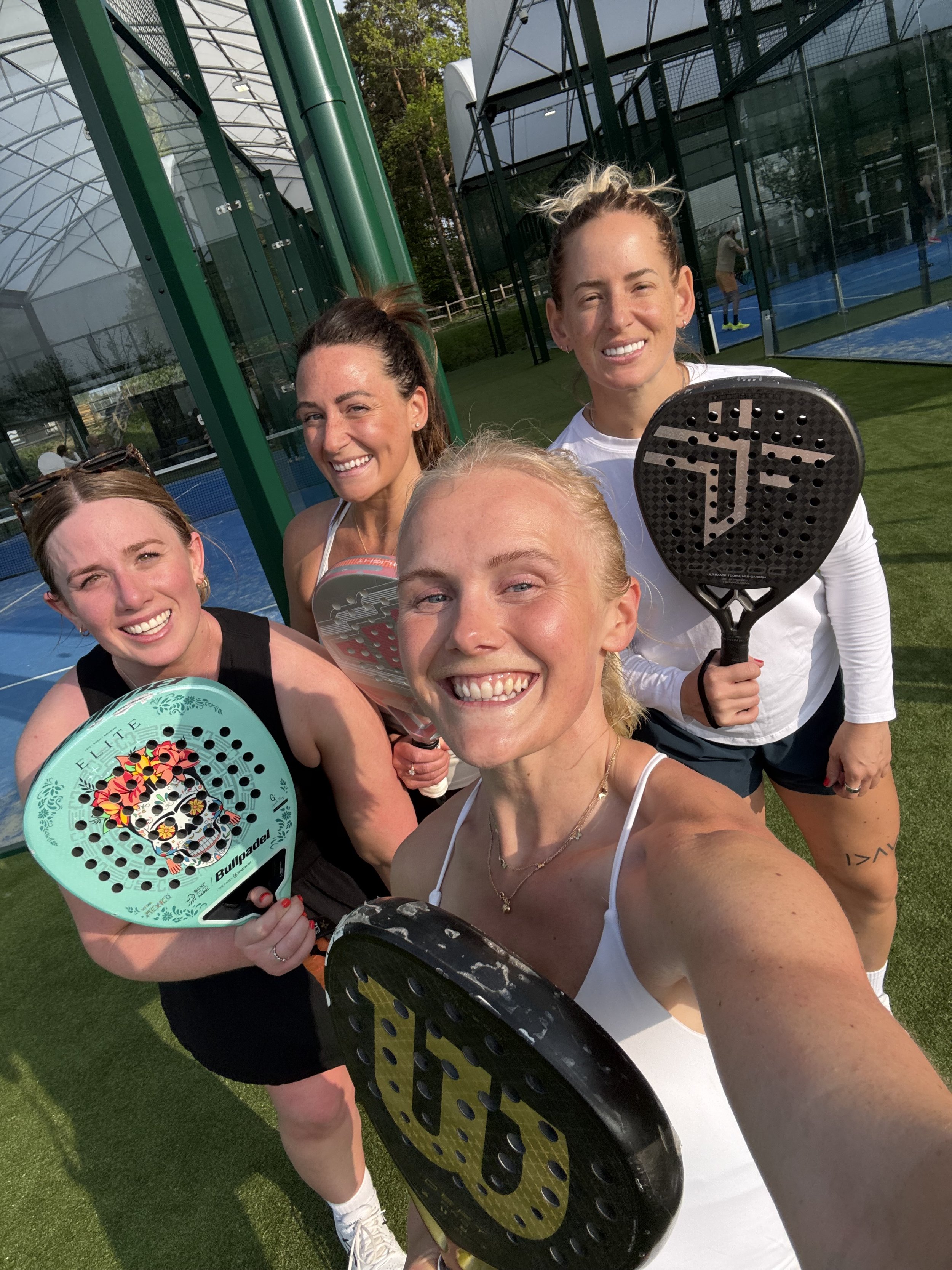 Group of four women holding padel rackets on outdoor tennis court, smiling, with glass-walled sports facilities in the background.