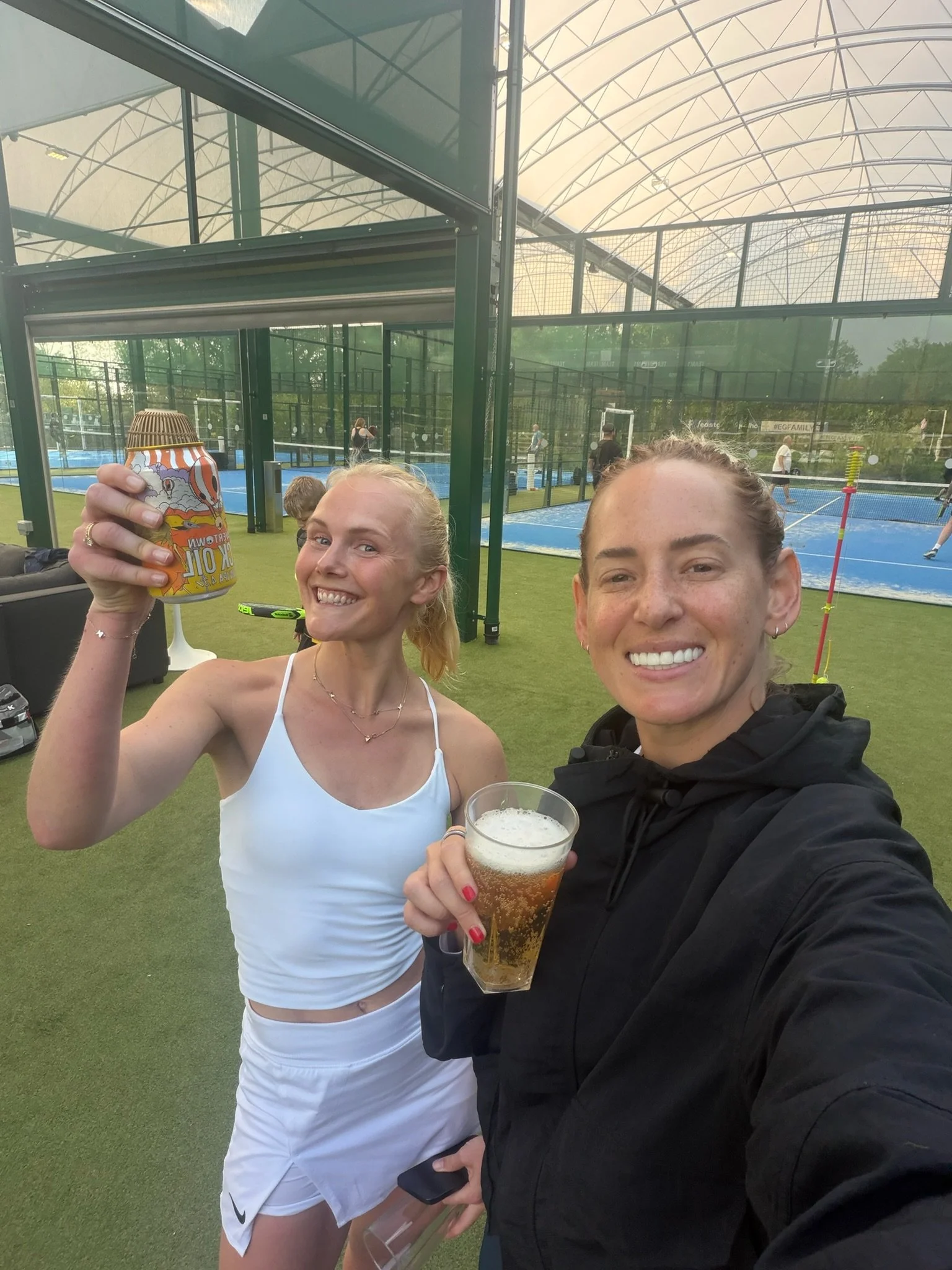Two women smiling and holding drinks, one with a can of beer and the other with a glass of beer, at an indoor tennis court facility with players practicing in the background.