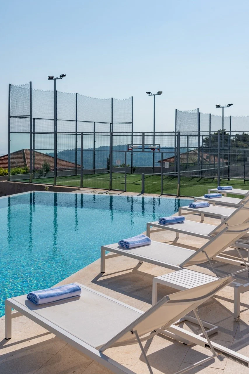 Poolside view with white lounge chairs and blue towels, tennis court with tall fencing, and scenic hills in the background.