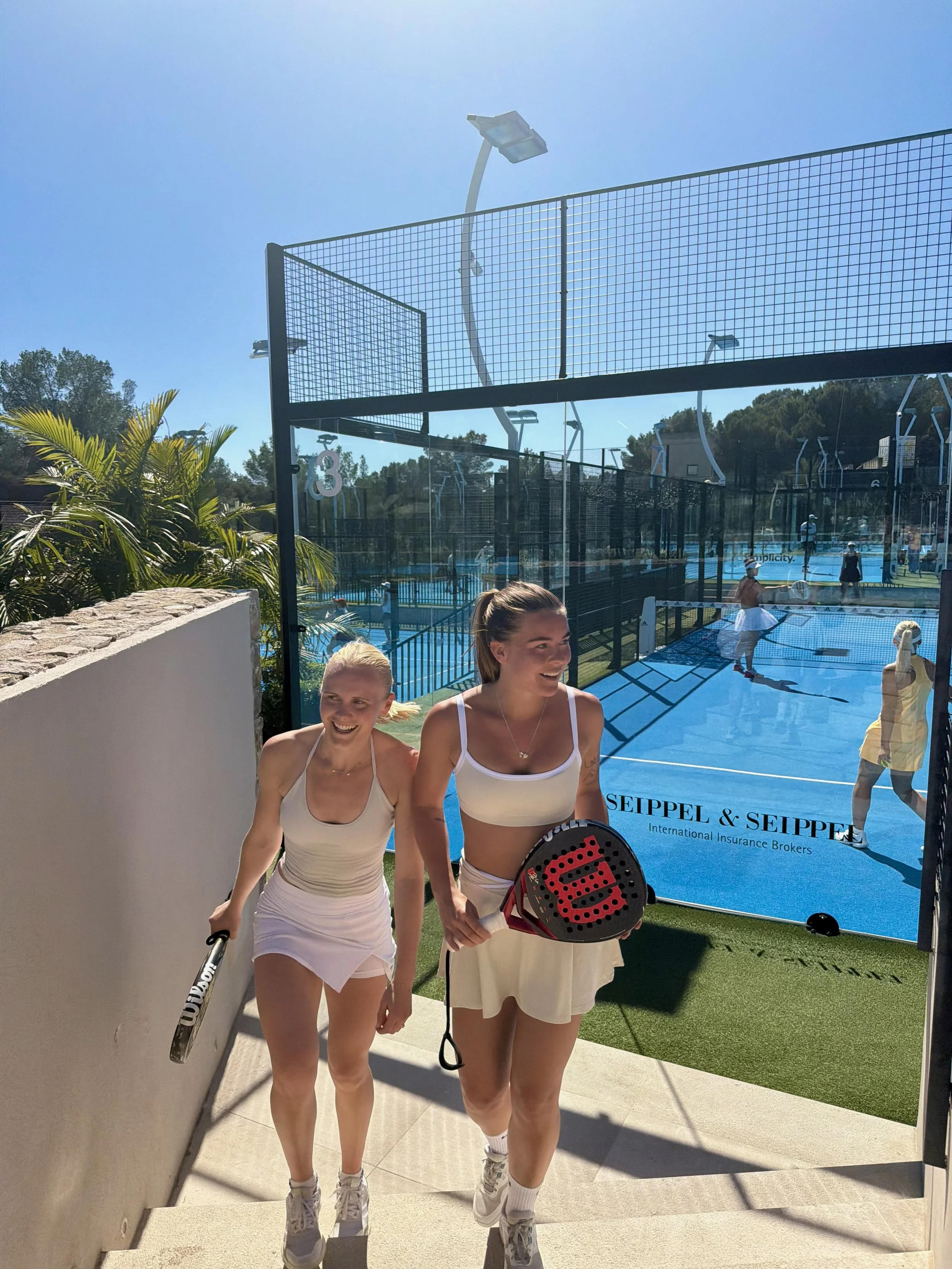 Two young women in tennis outfits walking through a glass door at a tennis facility, with paddle tennis courts and a blue sign with black text in the background.