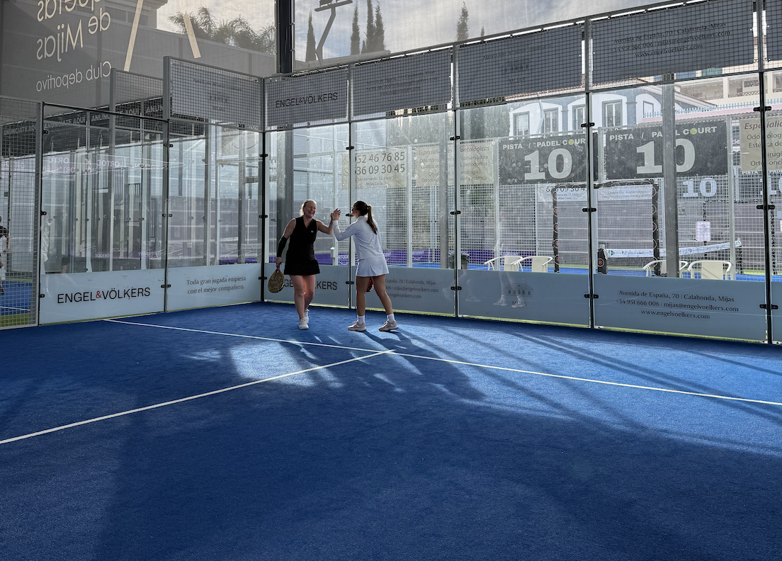 Two women playing padel on an indoor court, high-fiving each other. One woman is dressed in black, holding a paddle, and the other in white. The court has a blue surface and glass walls with signage.