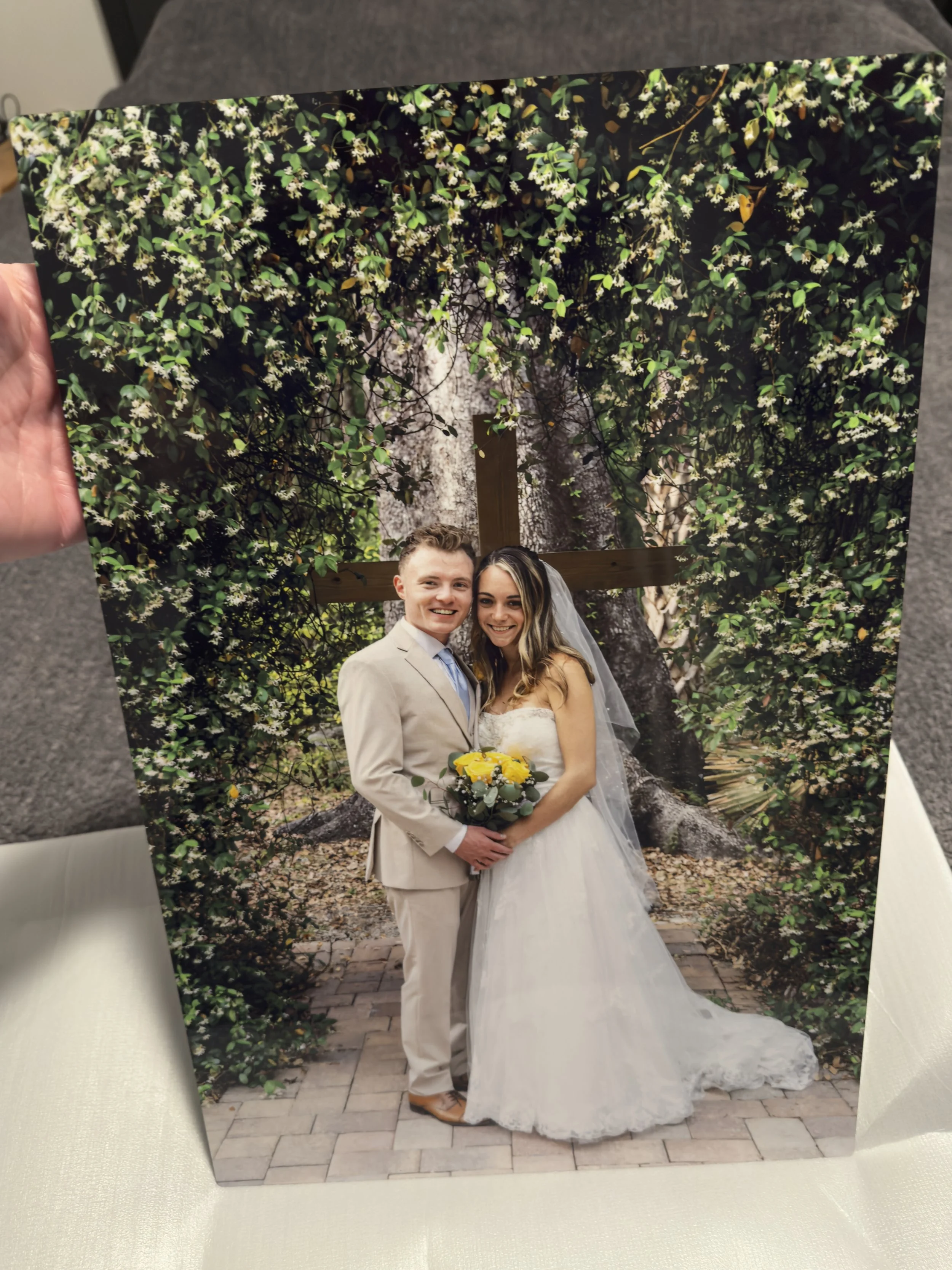 A wedding photo of a bride and groom standing together outdoors in front of a large tree with greenery and white flowers, holding a bouquet of yellow flowers.