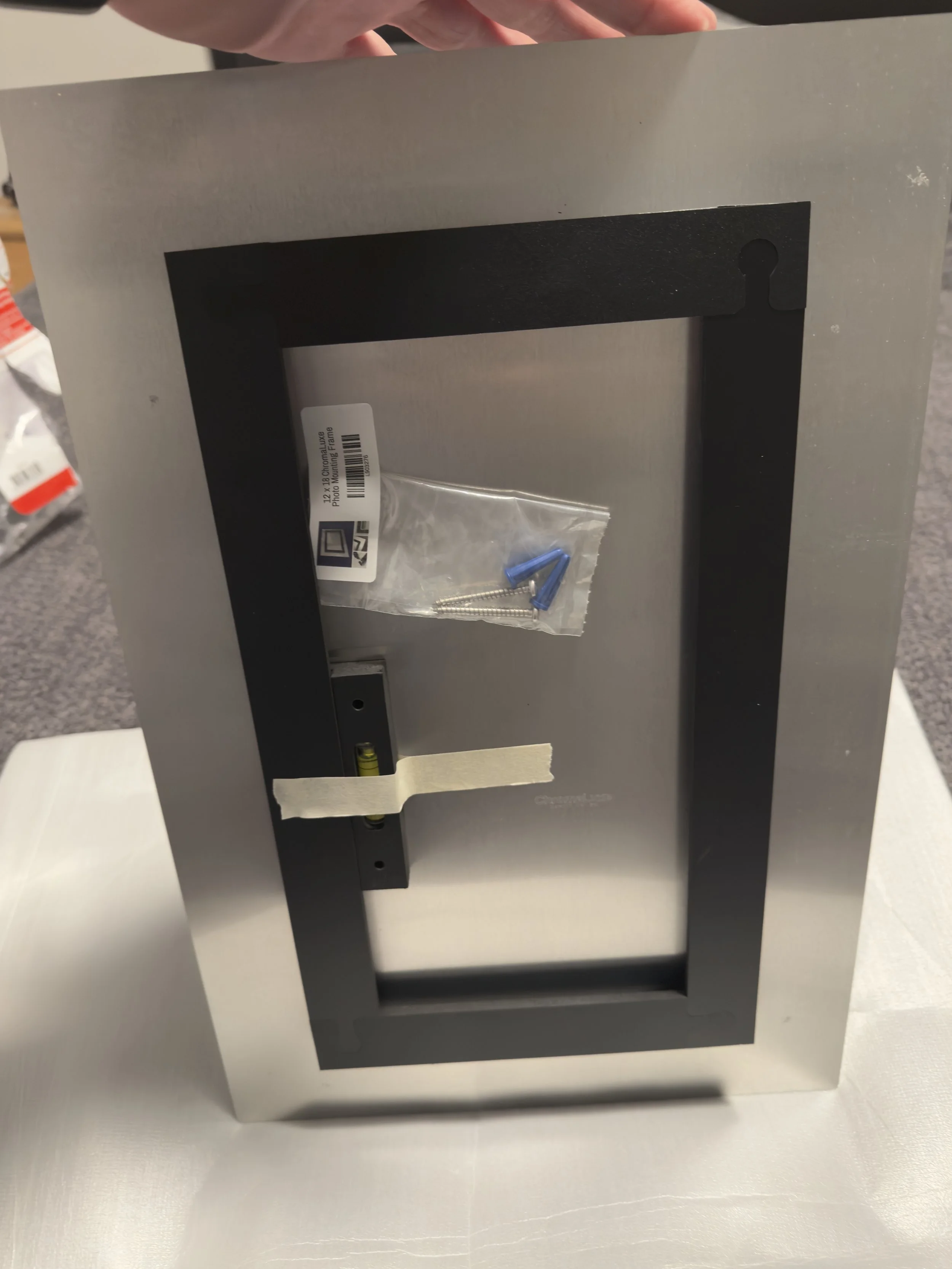 A metallic electrical box frame with a small plastic bag containing screws and blue anchors, and a bubble level attached with tape.