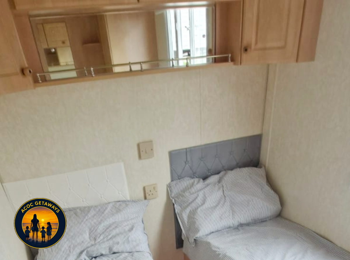Two beds with striped linens in a small room with beige wallpaper, electrical outlets and a wooden shelf with a mirror. A reflection in the mirror shows a wooden door and part of a window.