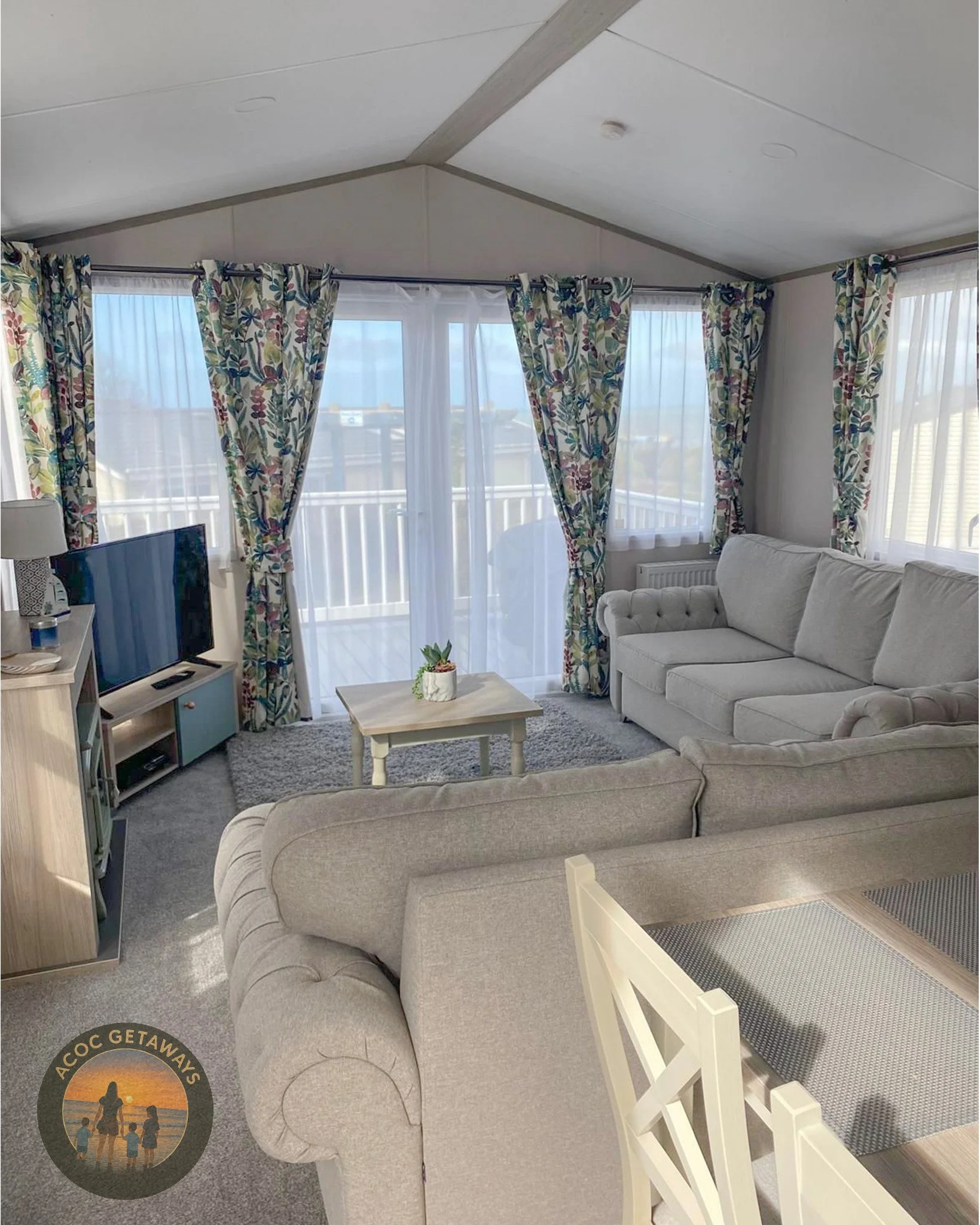 Living room with beige sofas, a small coffee table with a potted plant, a TV on a stand, and large windows with floral curtains and sheer white drapes leading to a balcony with ocean view.