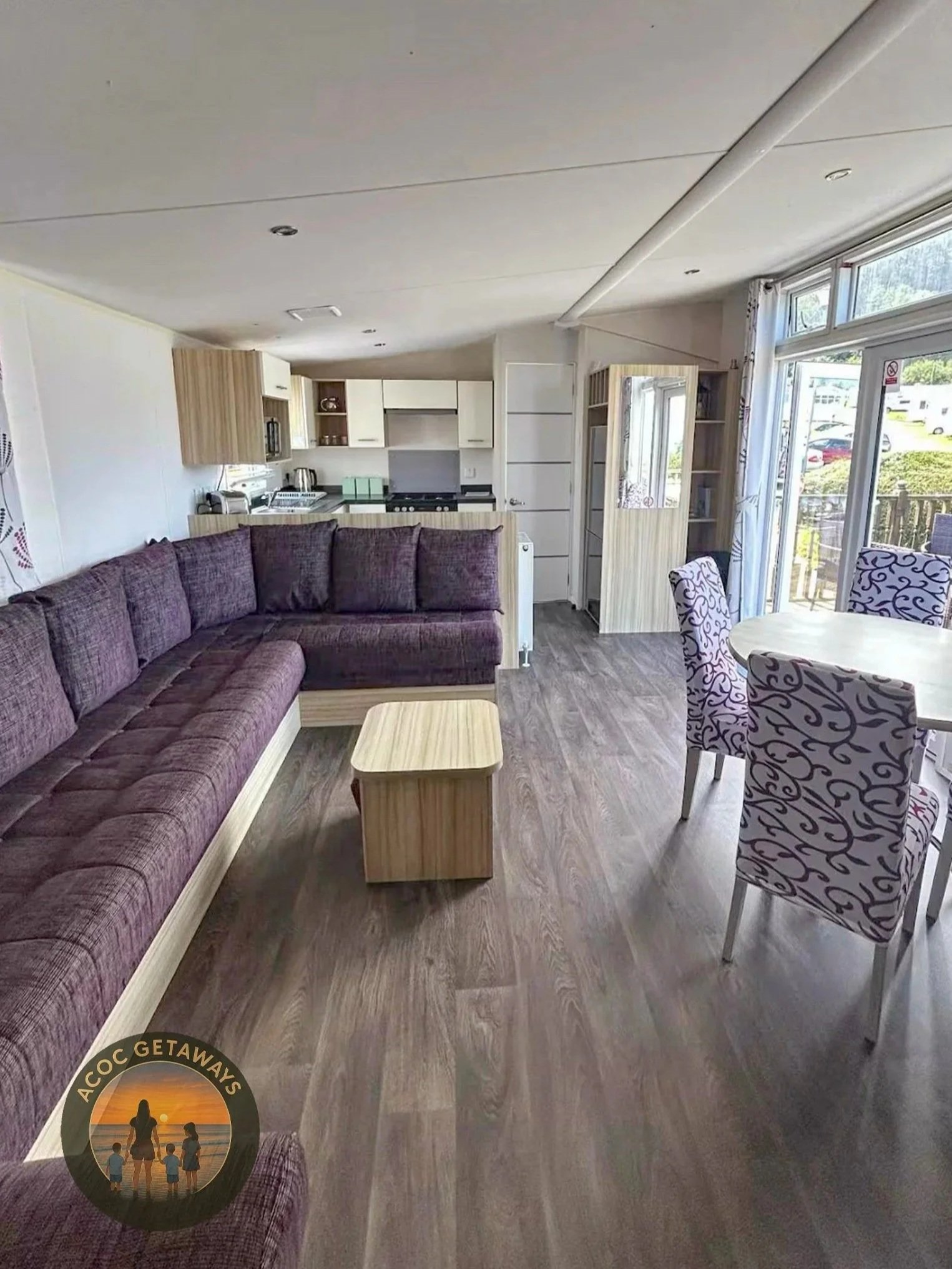 Living room with purple sectional sofa, wooden coffee table, dining table with patterned chairs, open kitchen with white cabinets, and large windows with sliding glass door leading outside.