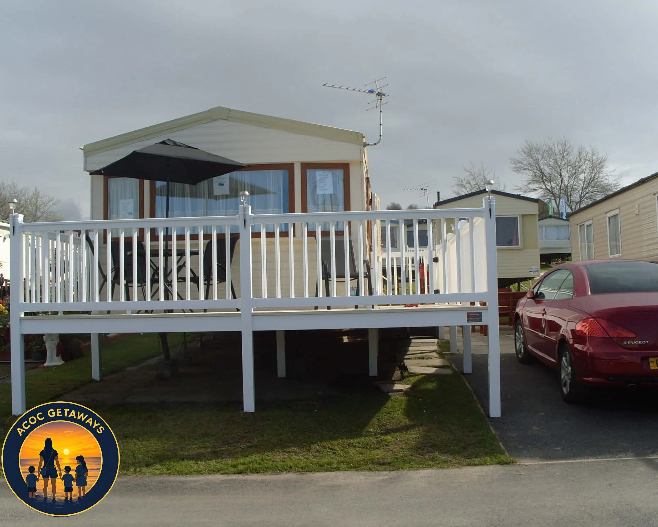 A mobile home with a white railing deck, outdoor furniture including a table, chairs, and a patio umbrella, parked next to a red car on the driveway, under a cloudy sky.