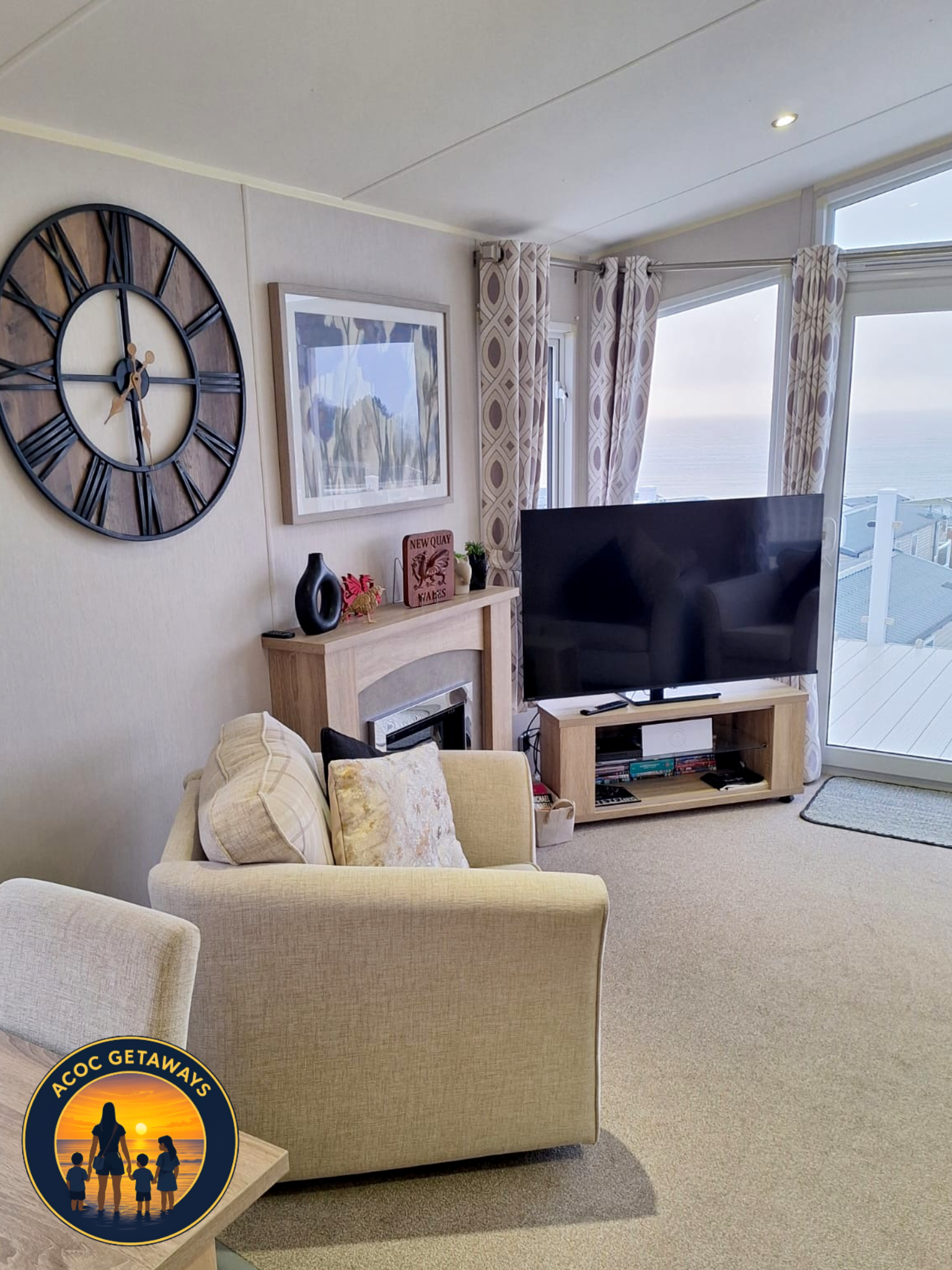 Living room with a cream sofa, wall clock, framed artwork, TV on a stand, and large ocean-view windows with patterned curtains.