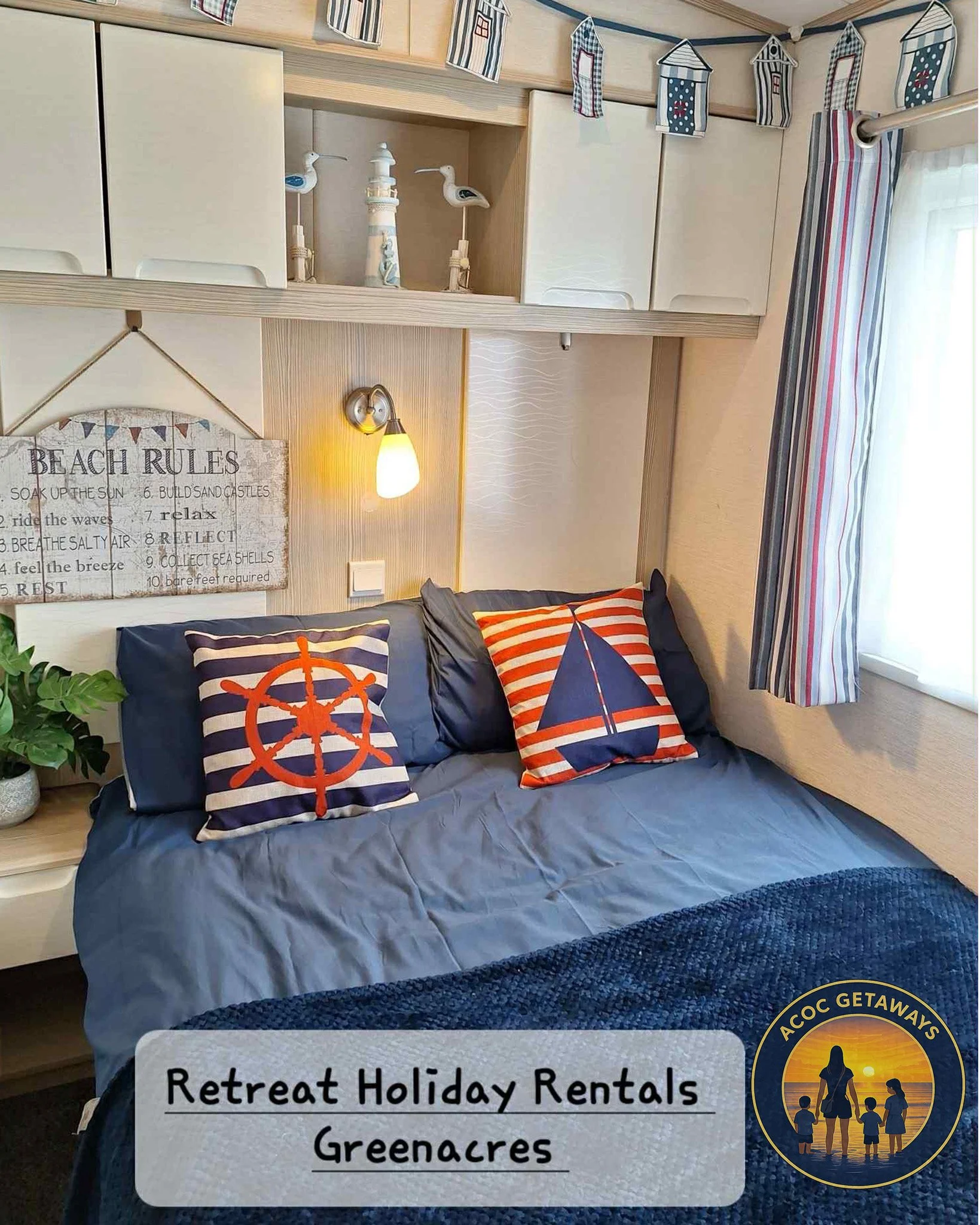 A cozy bedroom with a bed featuring two decorative pillows with nautical designs, colorful striped curtains, a wall-mounted lamp, and a decorative banner of houses hanging from the ceiling. There is a beach rules sign on the wall, and a small plant i