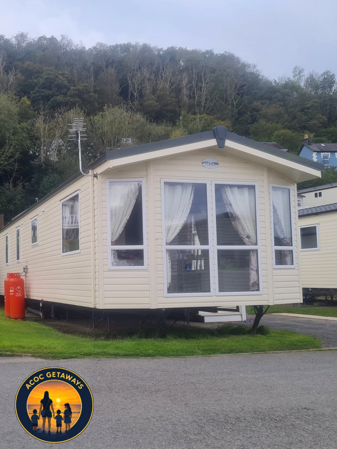 A beige mobile home with large windows and white curtains, situated on a grassy area with a hill covered in trees in the background. A red gas tank is visible on the left side of the mobile home. The logo 'ACOC GETAWAYS' featuring a family silhouette