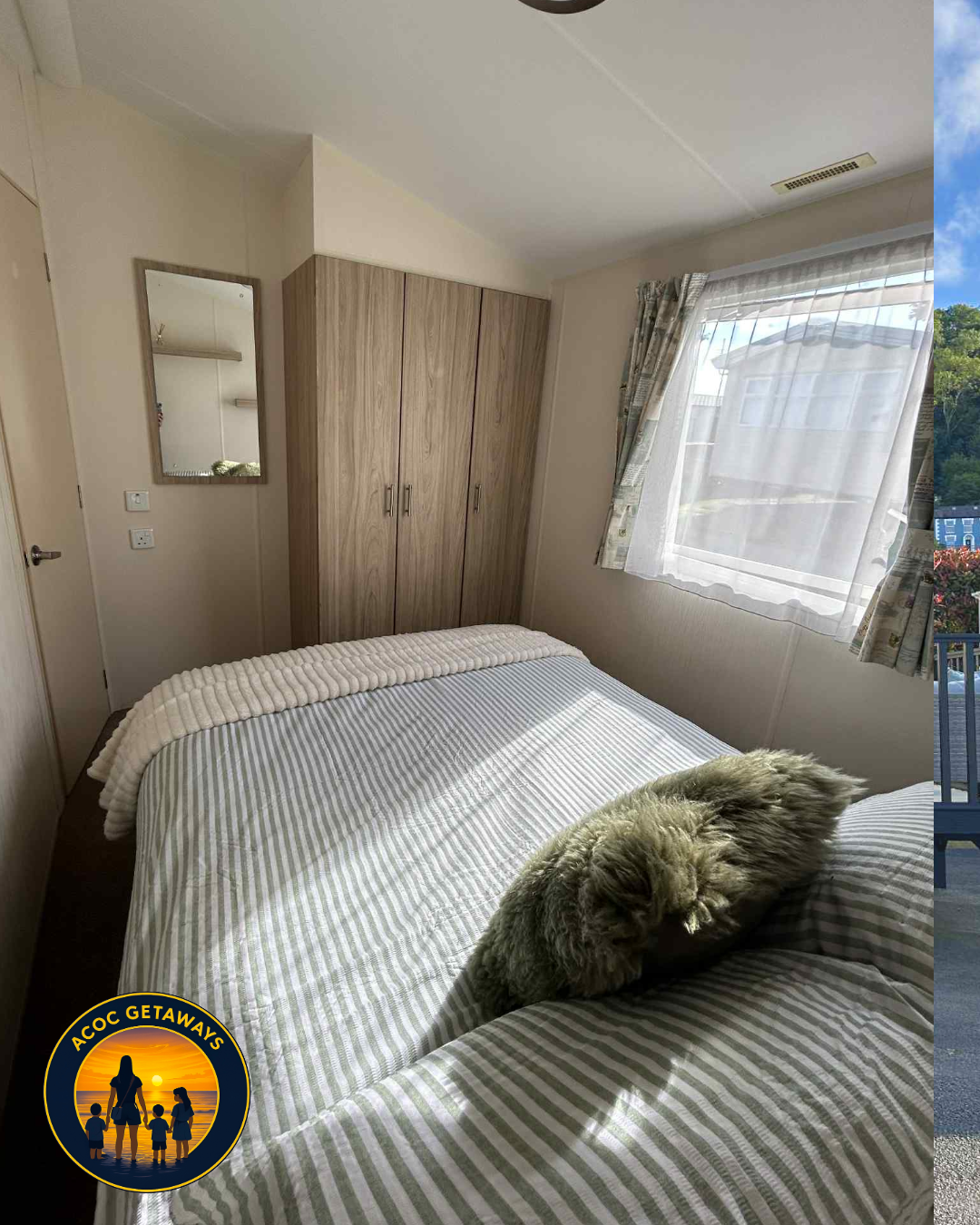 A cozy bedroom with a bed covered in a striped bedspread, a furry throw pillow, and a sleeping gray fluffy cat. There is a wooden wardrobe, a wall mirror, a window with sheer and patterned curtains allowing sunlight inside, and a small outdoor balcon