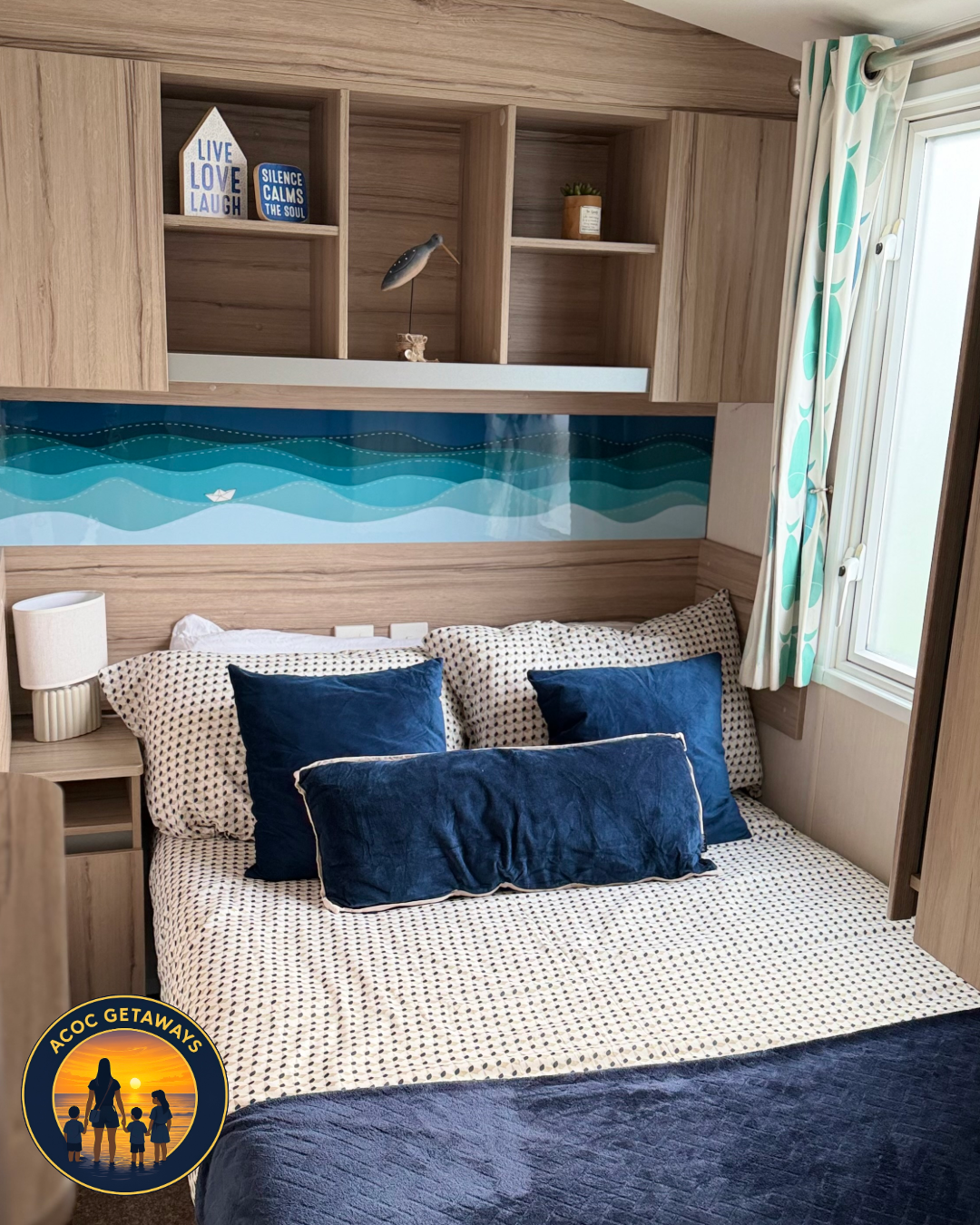 A cozy bedroom with a wooden headboard and built-in shelves above the bed, decorated with a sea-themed mural, blue pillows, and patterned bedding. A window with patterned curtains is on the right side.