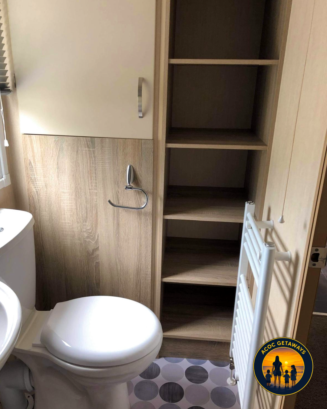 A small bathroom with a toilet, a wooden shelf, a small cabinet, a towel ring, a white towel radiator, and a window with blinds.