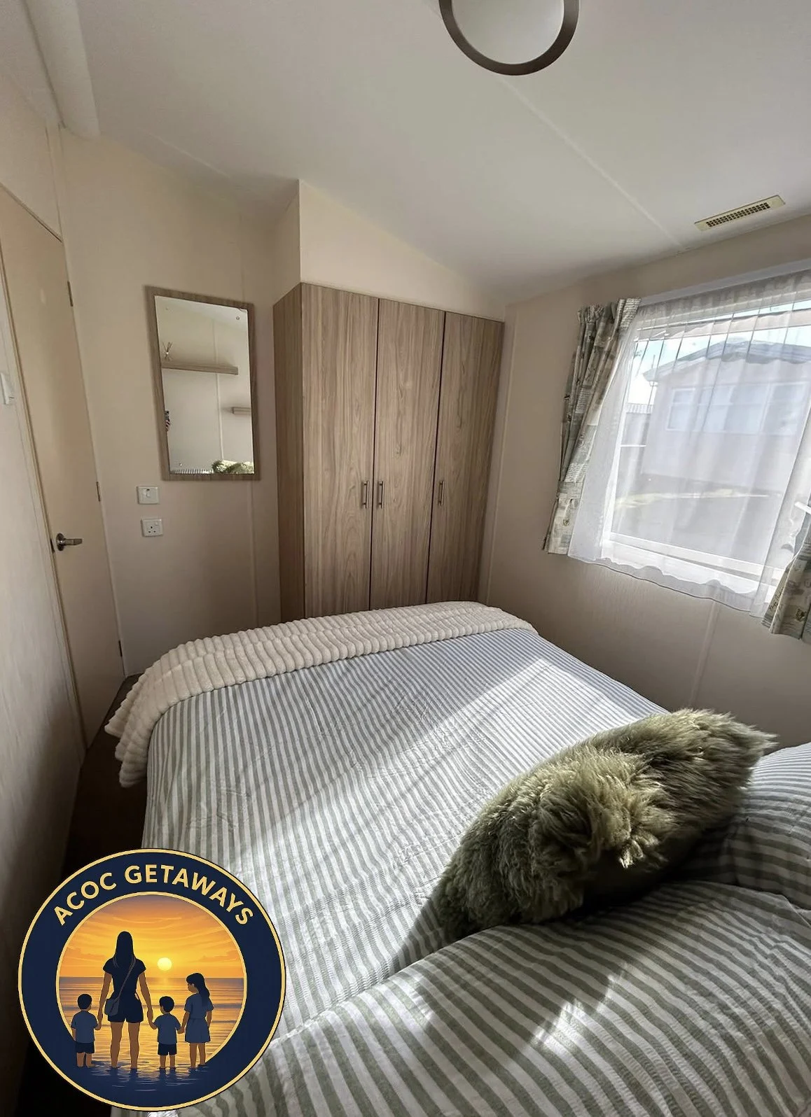 A cozy bedroom with a bed covered in striped linens, a fluffy blanket, and a sleeping cat. There is a wooden wardrobe, a large window with curtains, and a mirror on the wall.