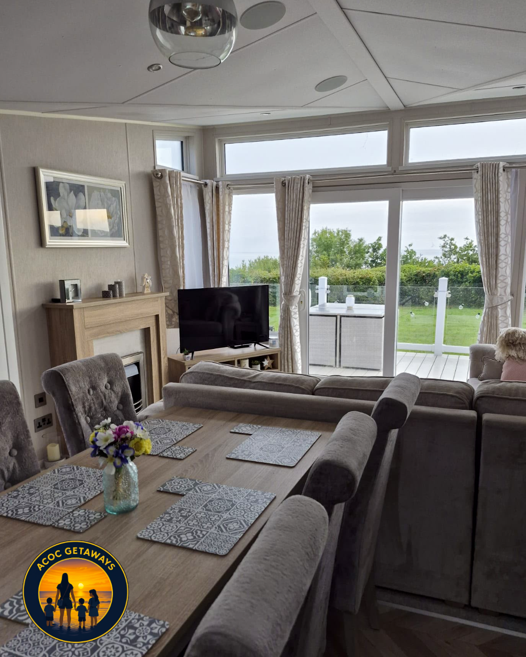 Living room with large windows and a sliding glass door leading to a patio with a green lawn. There is a dining table with floral centerpiece and placemats, a gray sectional sofa, a TV on a stand, a fireplace, and framed artwork on the wall.