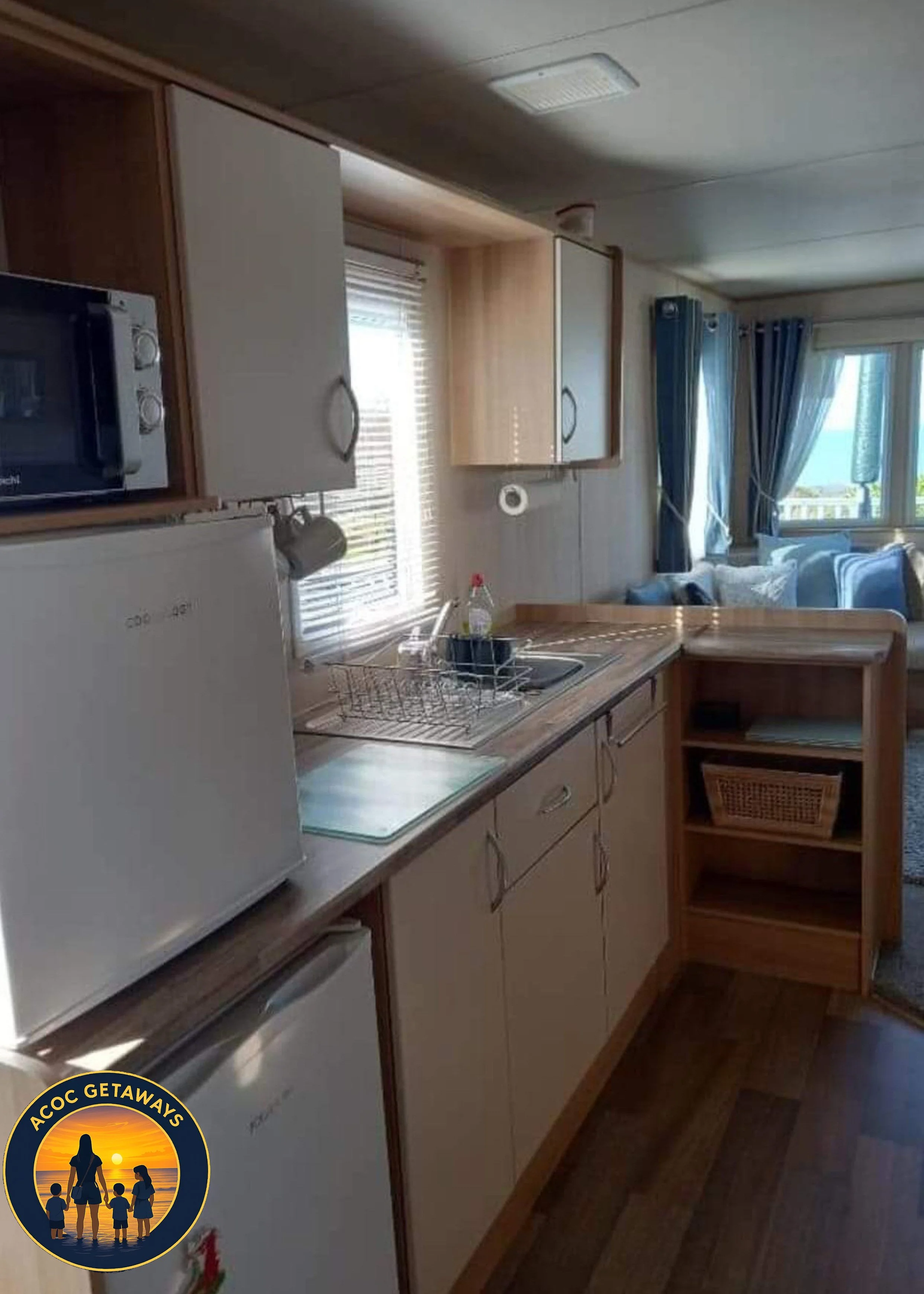 Small kitchen with white refrigerator, microwave, sink, and wooden cabinets. Open layout with view of living room and large window with blue curtains and ocean view.