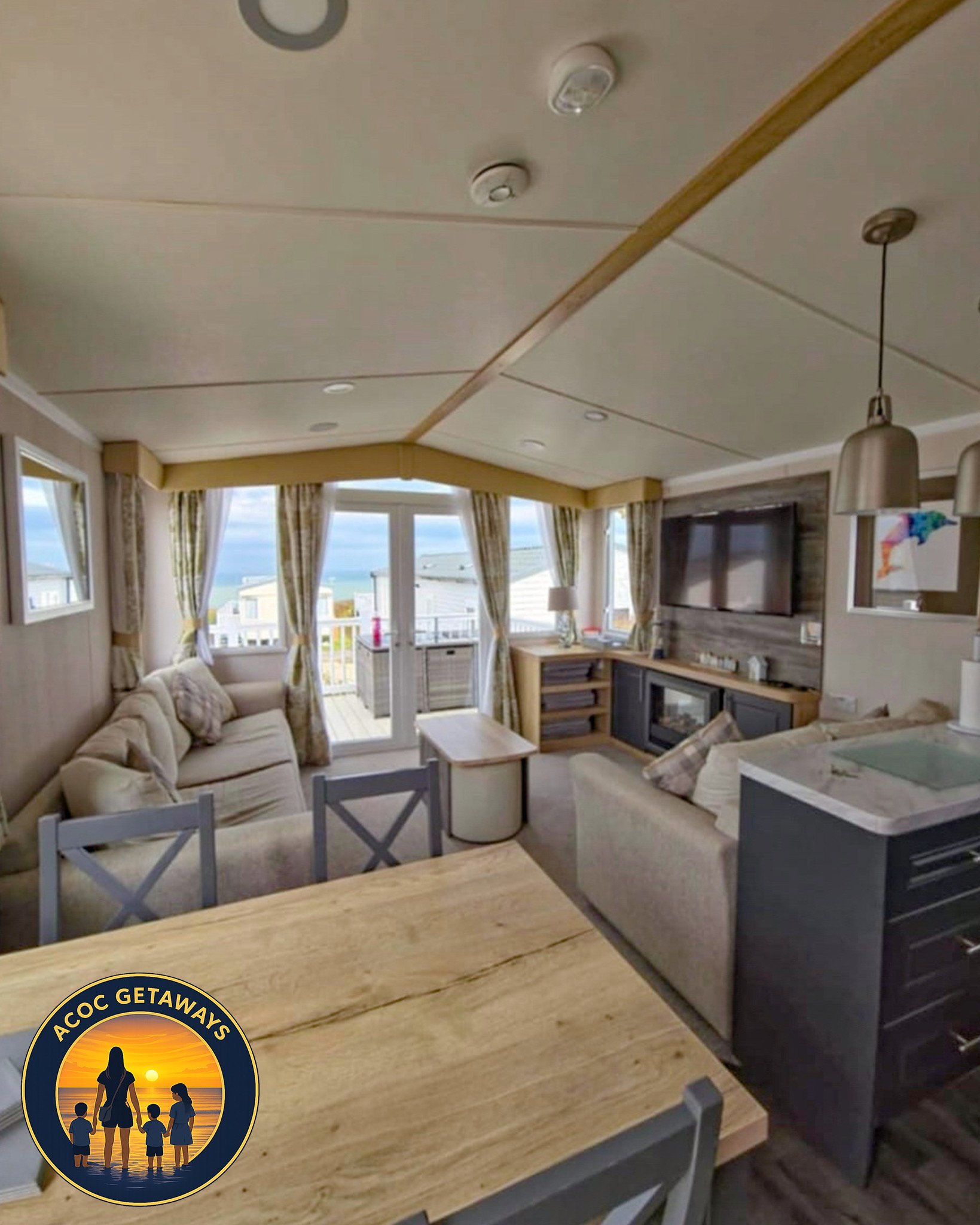 A cozy living room with large windows revealing a view of neighboring houses and the ocean. The room features two beige sofas, a wooden dining table with gray chairs, a flat-screen TV, and a fireplace. A balcony is accessible through a glass door, an