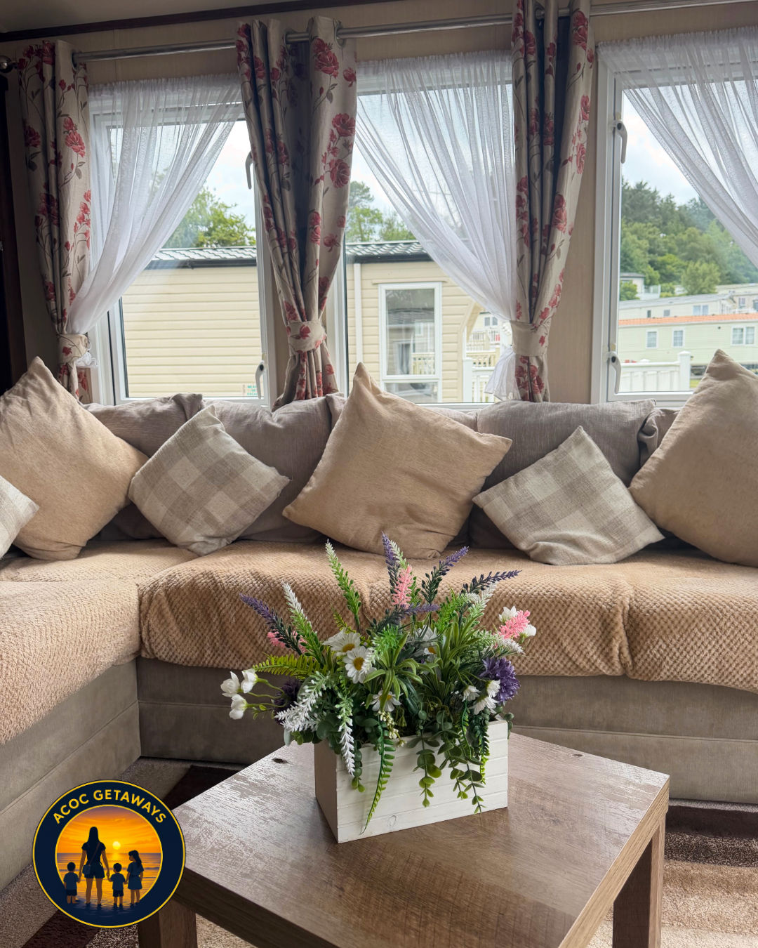 A cozy living room with a beige L-shaped sofa adorned with various beige and checkered pillows, in front of a window with floral curtains. A wooden coffee table with a white flower arrangement is positioned in front of the sofa, with a background of 