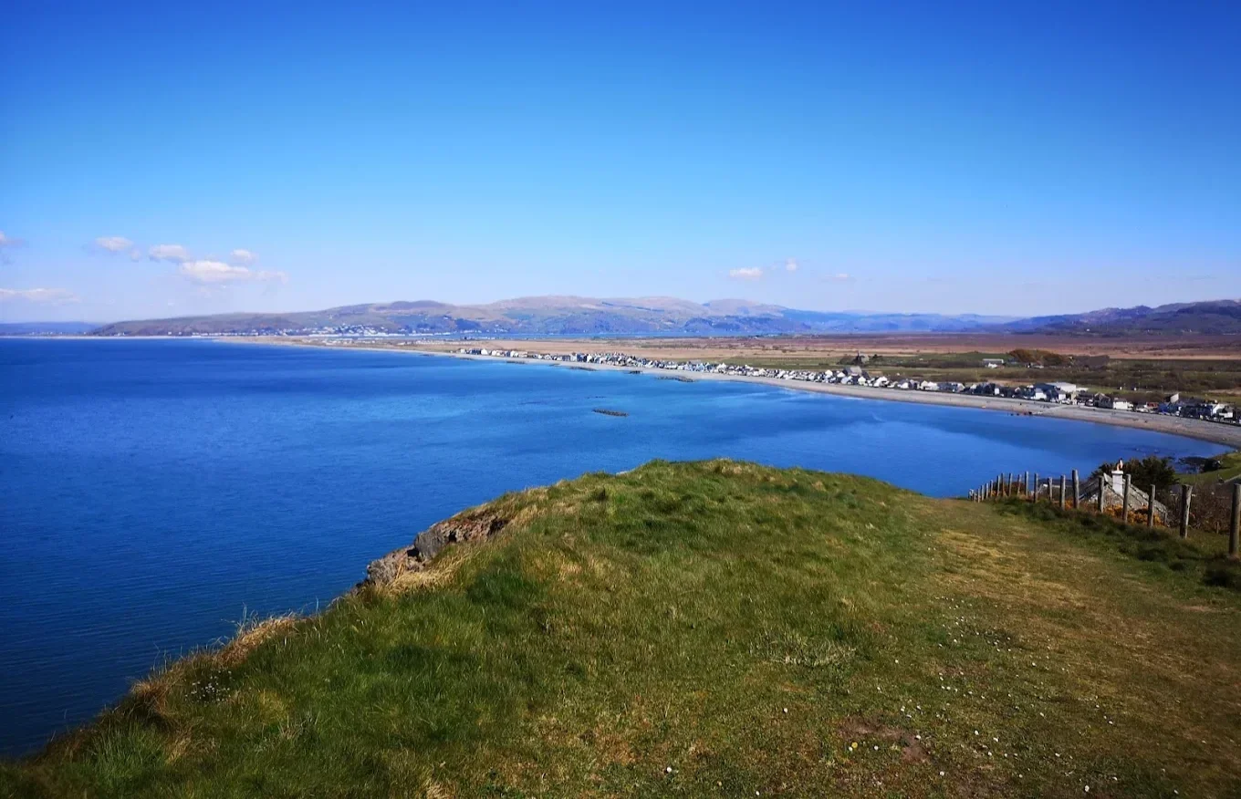 A scenic view of a coastline with a grassy hill in the foreground, a calm blue sea, a long beach with houses along the shore, and distant rolling hills under a clear blue sky.