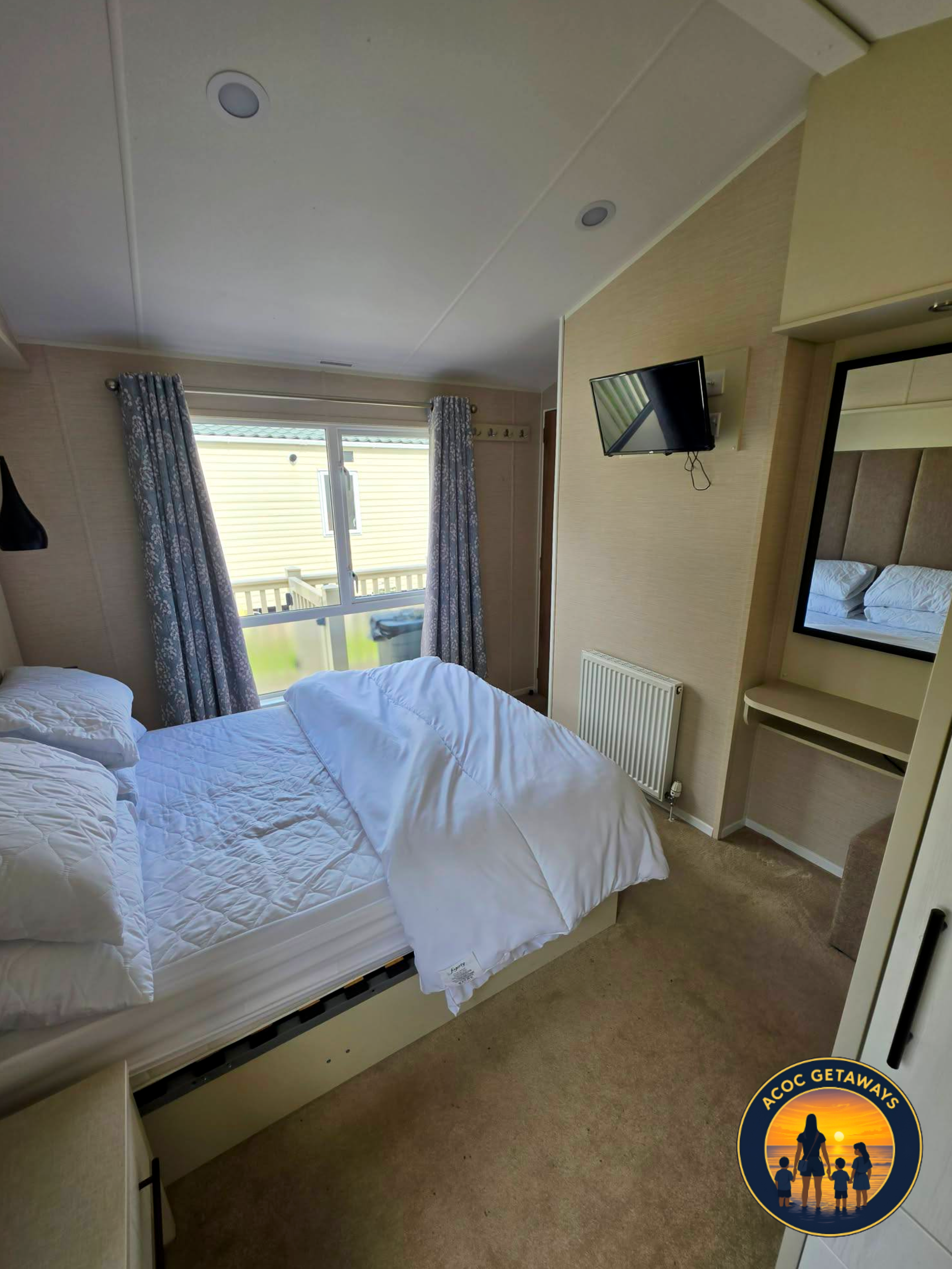 View of a bedroom with a bed, window with curtains, a small mirror, a wall-mounted TV, and a radiator.