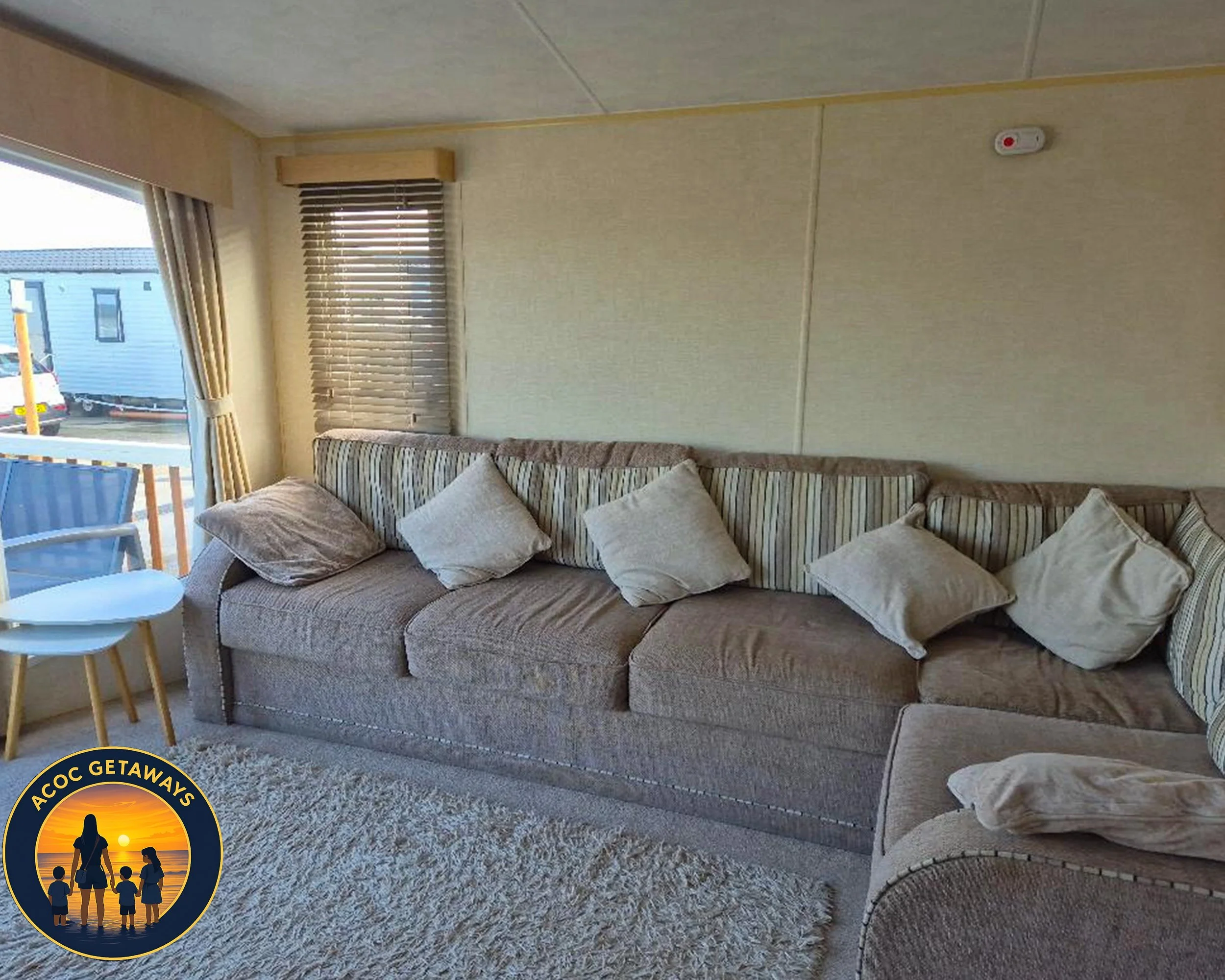 Living room with a beige, patterned sectional sofa, several light-colored pillows, a fluffy rug, and a window with curtains and blinds. There's an outdoor area visible through the window.