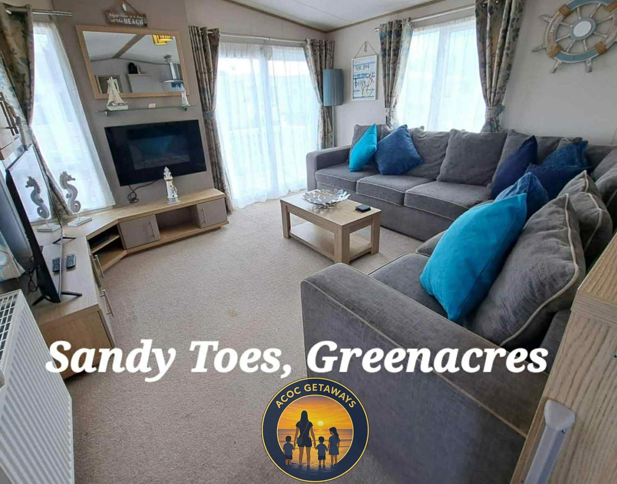 Living room with gray sofas decorated with blue pillows, wooden coffee table, large windows with floral curtains, TV mounted on the wall, and beach-themed decorations, including seahorse figures and a ship wheel, with sunlight streaming through the curtains.