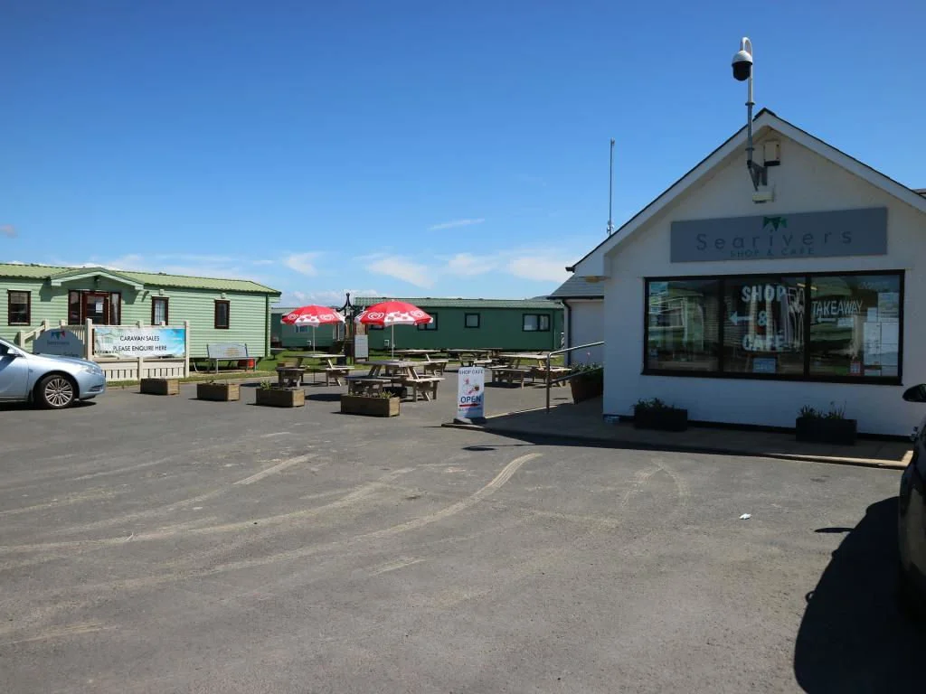 Outdoor view of Seabird's shop and takeaway with a parking lot, picnic tables, umbrellas, and a caravan sales sign, under a clear blue sky.