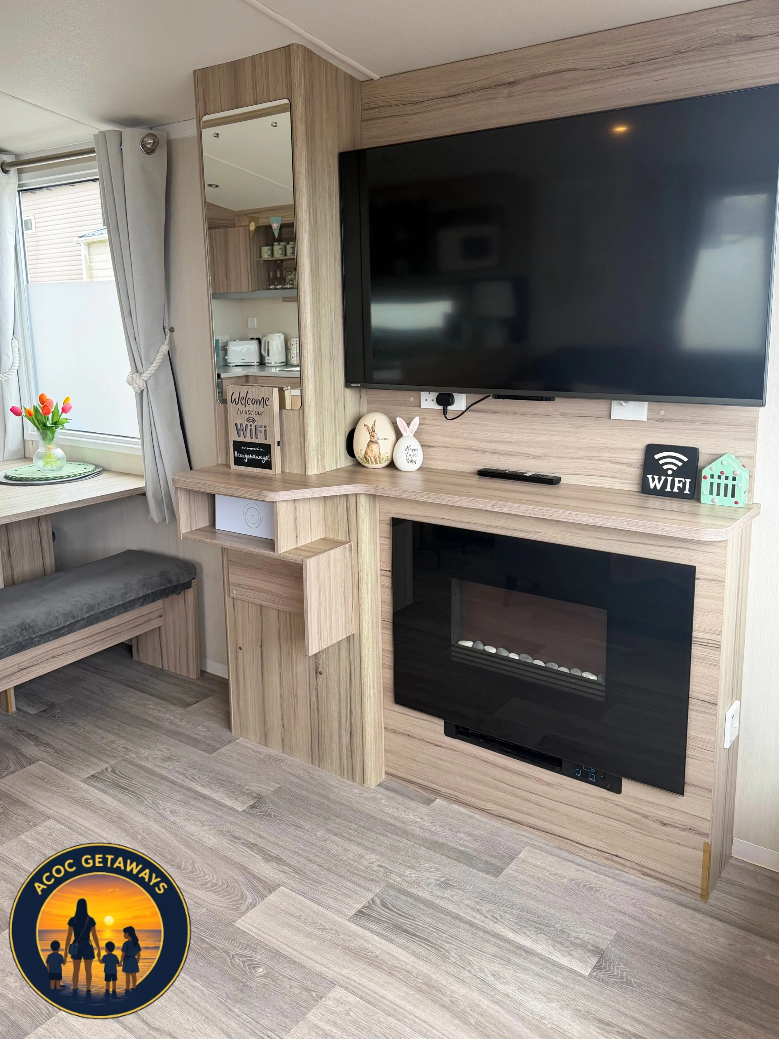 Living room with a mounted flat-screen TV, a decorative wood panel, a mirror, and a small shelf with bunny and egg decorations. A corner of a window with curtains and a small table with tulips in a vase are visible.