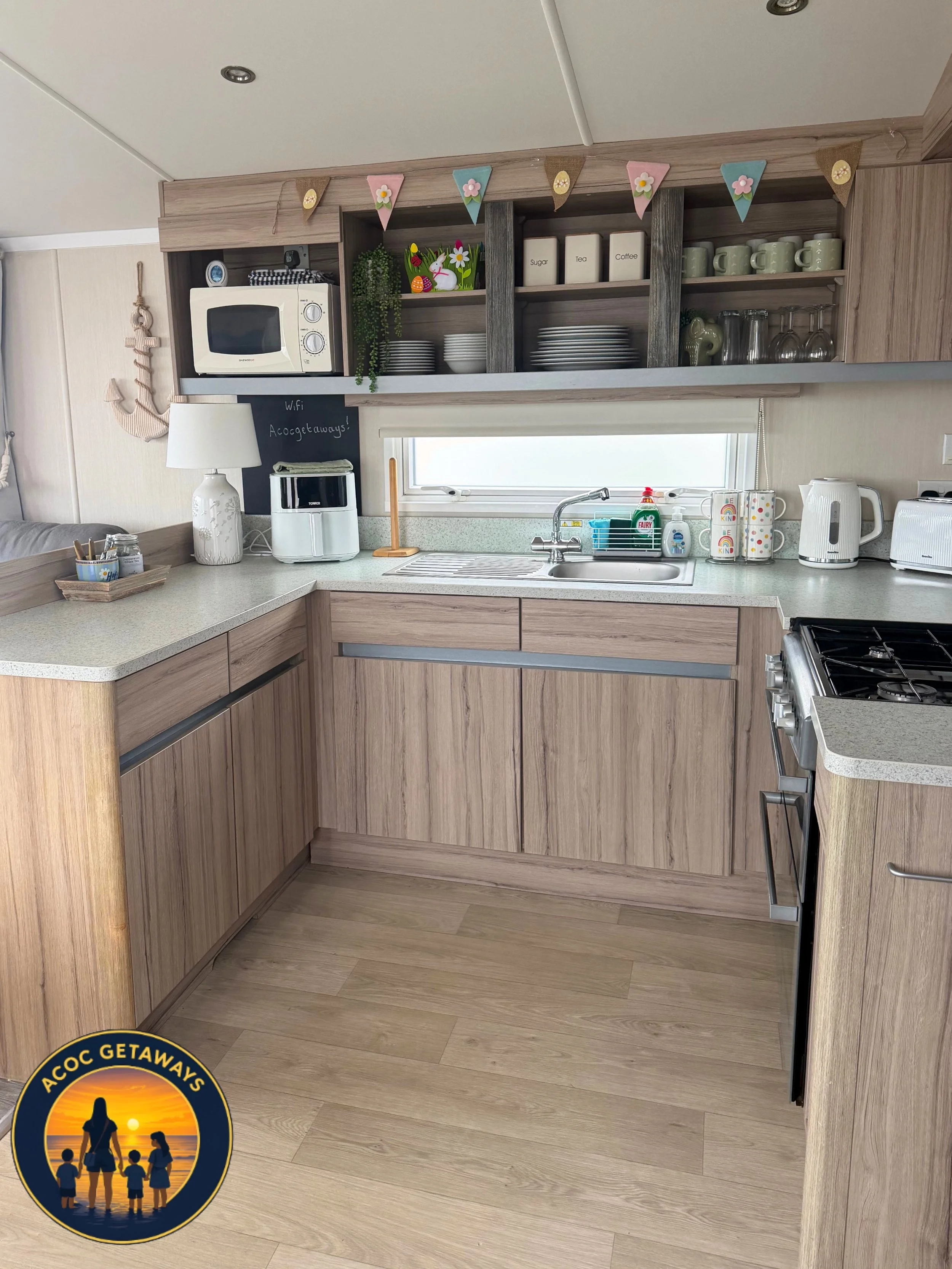 Kitchen with light wood cabinets, beige countertop, white microwave, sink, and various kitchen items. Decor includes a string of pastel-colored flags with flower and egg accents, a small chalkboard sign saying "WiFi Acceptaways!", and a round logo wi