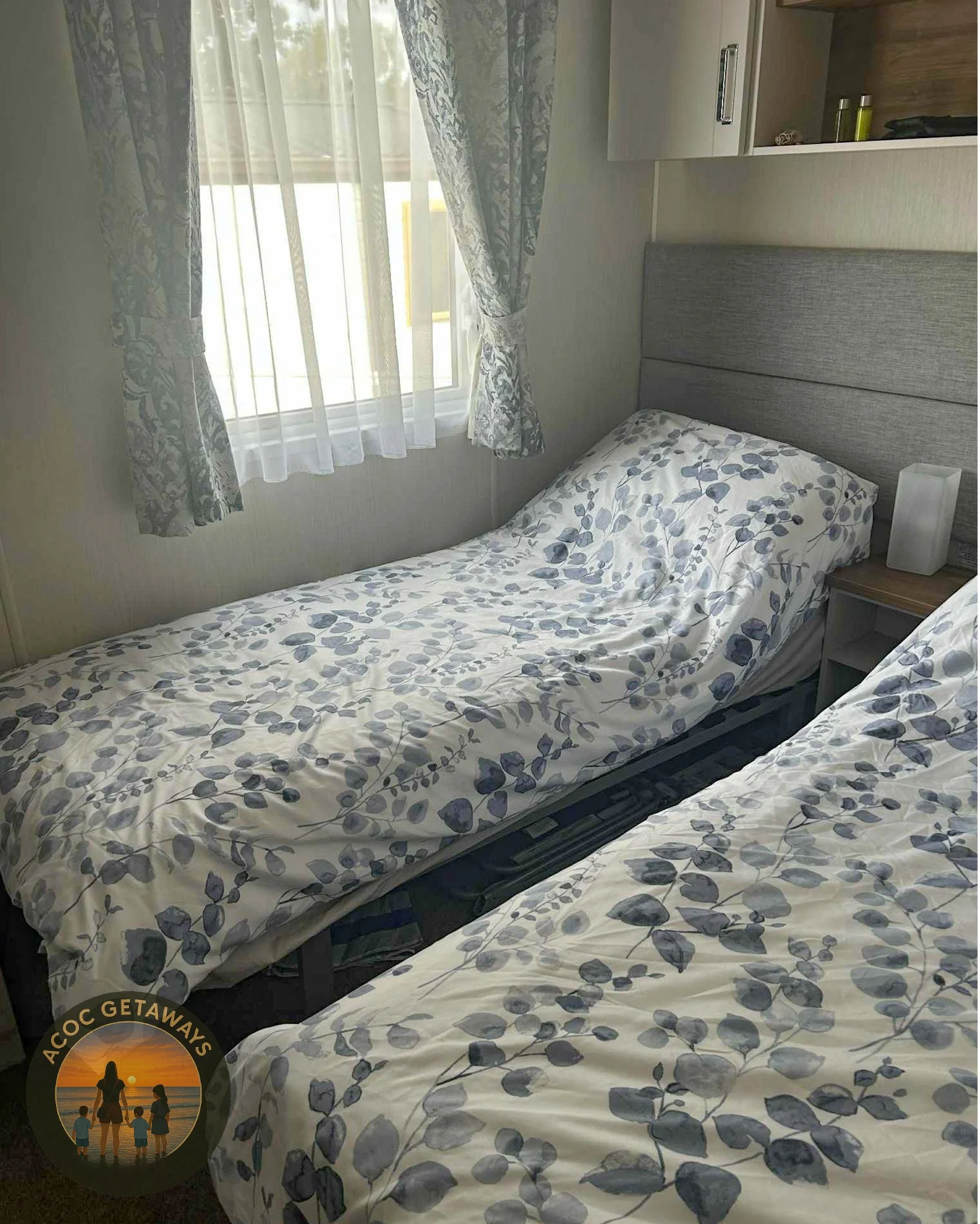 Two single beds in a bedroom with matching floral bedspreads, a window with lace curtains, and a wall-mounted cabinet with toiletries.