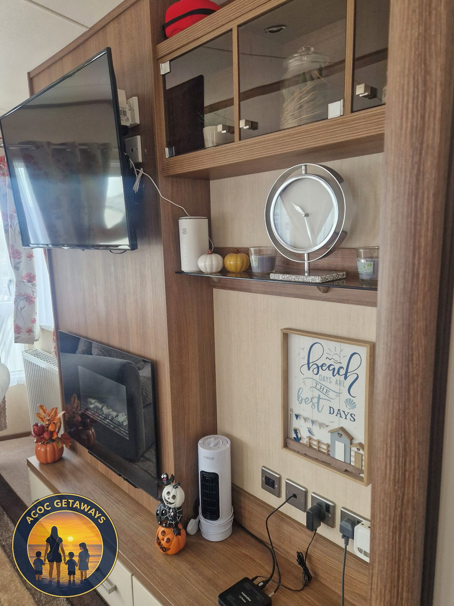 Wooden entertainment center with a flat-screen TV, decorative ghost and pumpkin figures, a clock, candles, and a framed beach-themed quote, in a cozy living room.