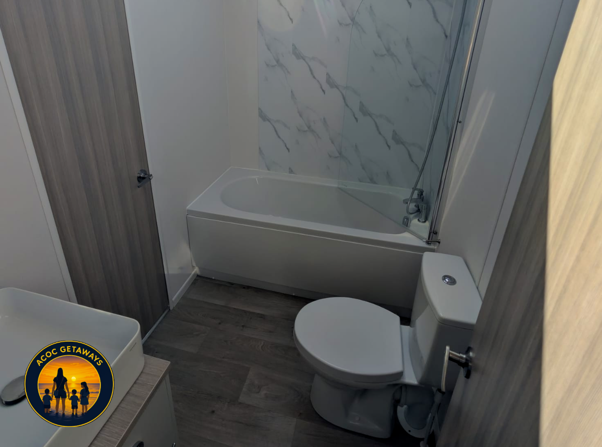 A small bathroom with a bathtub and a toilet. The bathtub has a marble-like wall behind it and a showerhead. There is a wooden door and a small countertop with a white sink and a black soap dish. The flooring is wood-like.
