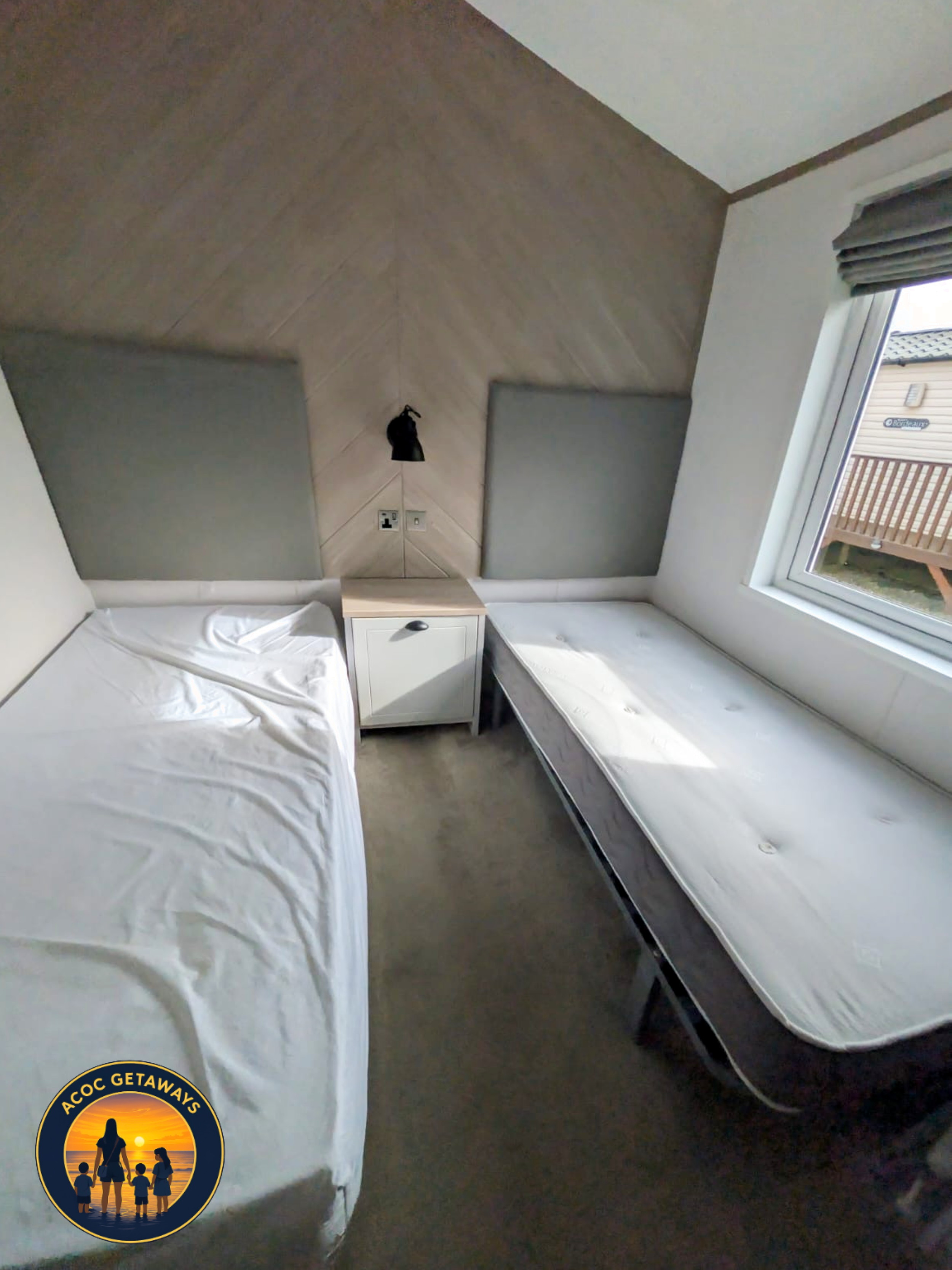 A bedroom with a bed covered in white sheets, a small white nightstand, gray soundproof panels on the wall, a window with gray blinds, and a view of a neighboring house's roof and balcony.
