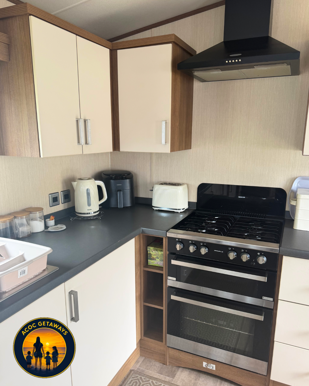 Kitchen with black countertop, white cabinets, a black stove, a black range hood, a white kettle, a black air fryer, a white toaster, small jars, a dish rack, and an 'A C O C GETAWAYS' logo with a sunset, silhouette of a woman and children.