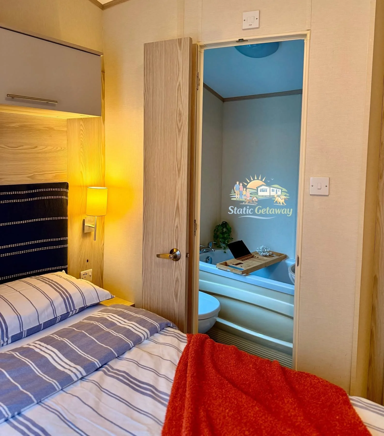 Bedroom with bed, striped bedding, bedside lamp, and a door open to a bathroom with a bathtub and wall art reading 'Static Getaway' featuring a sunset, lake, and cabin scene.