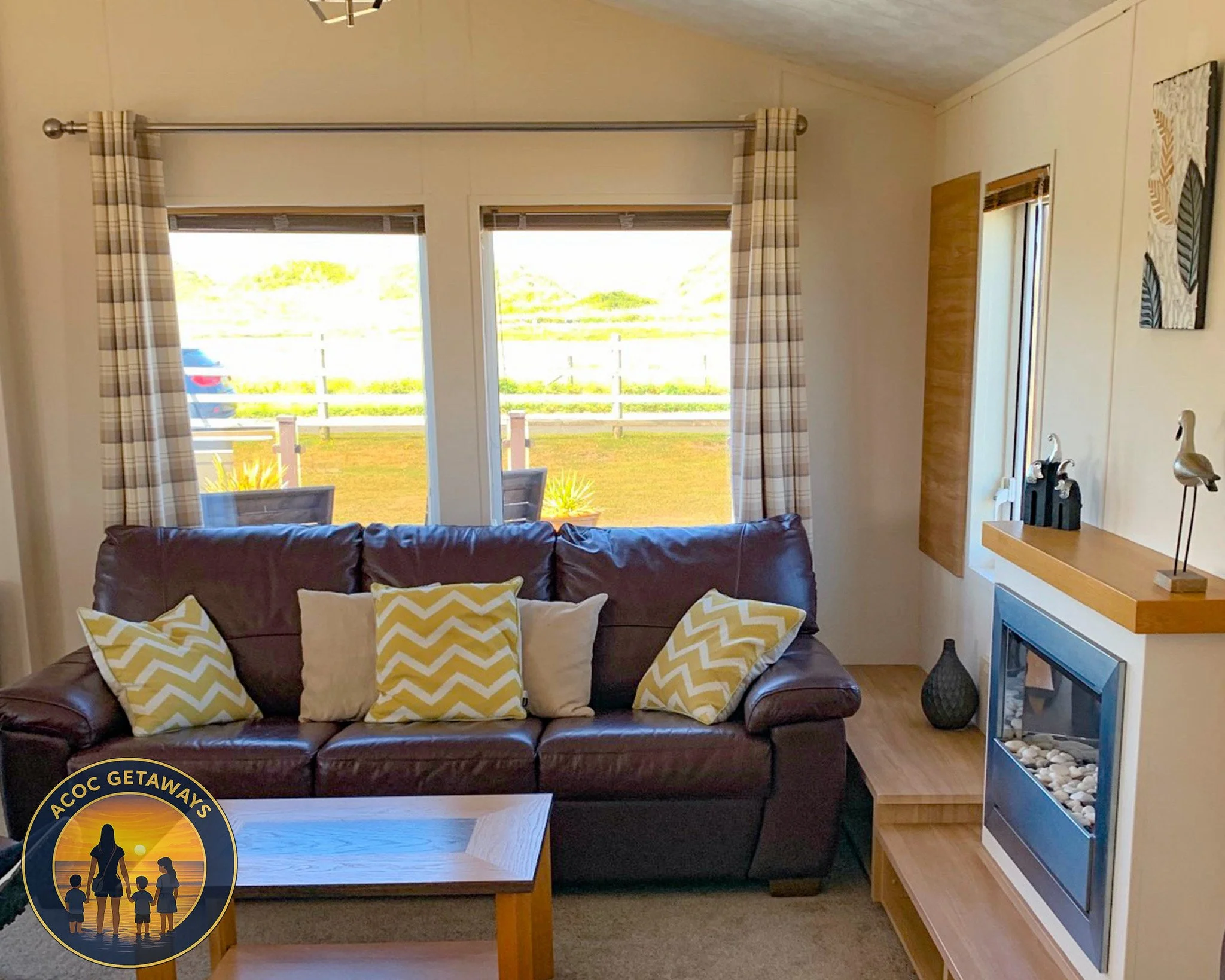 Living room with a brown leather sofa, yellow and white chevron-patterned pillows, a wooden coffee table, a fireplace with decorative stones, a wooden shelf with sailboat figurines, a window with plaid curtains showing a yard outside, and a wall hang