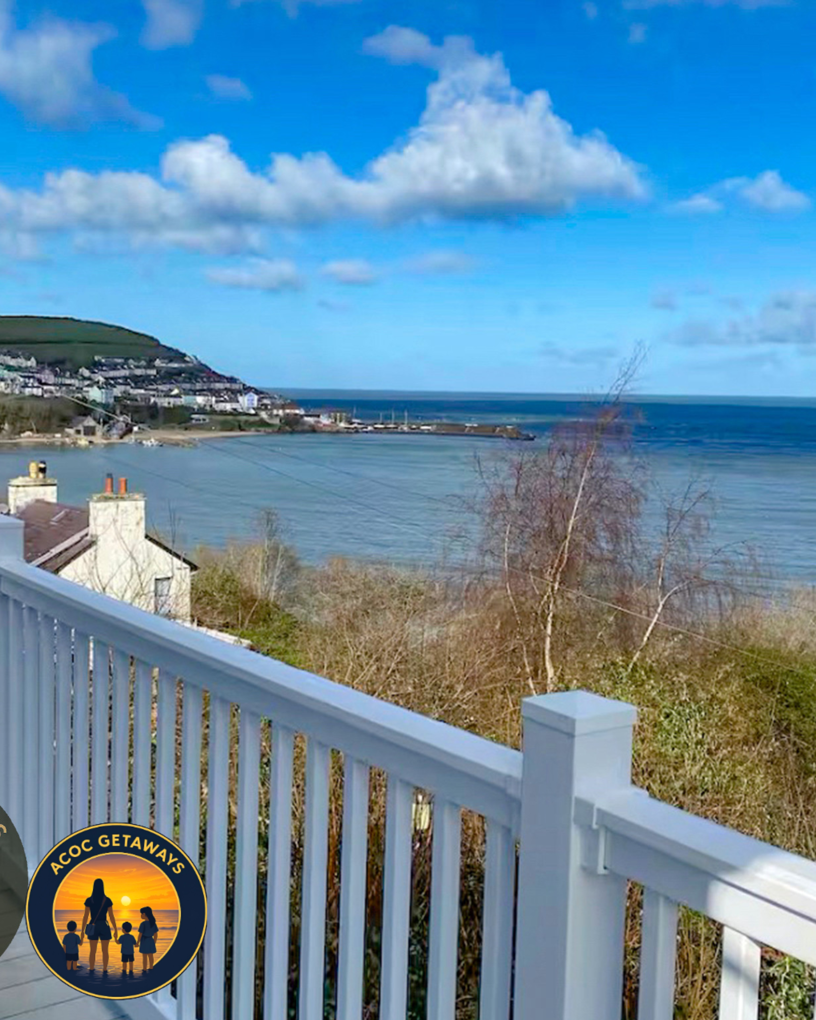 View of a seaside landscape with houses on a hill, blue sky with clouds, and ocean in the background. A white railing is in the foreground with a circular sticker featuring a sunset, silhouettes of a woman and children, and the text 'ACOC GETAWAYS'.