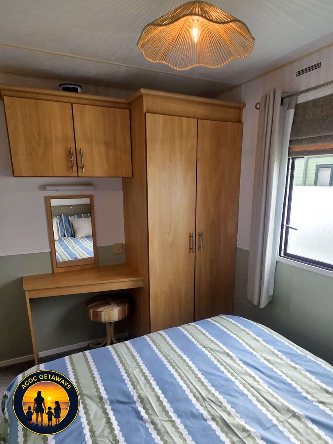 Bedroom with wooden wardrobe, bed with striped green, blue, and white bedding, small dressing table with mirror, and window with white curtains.