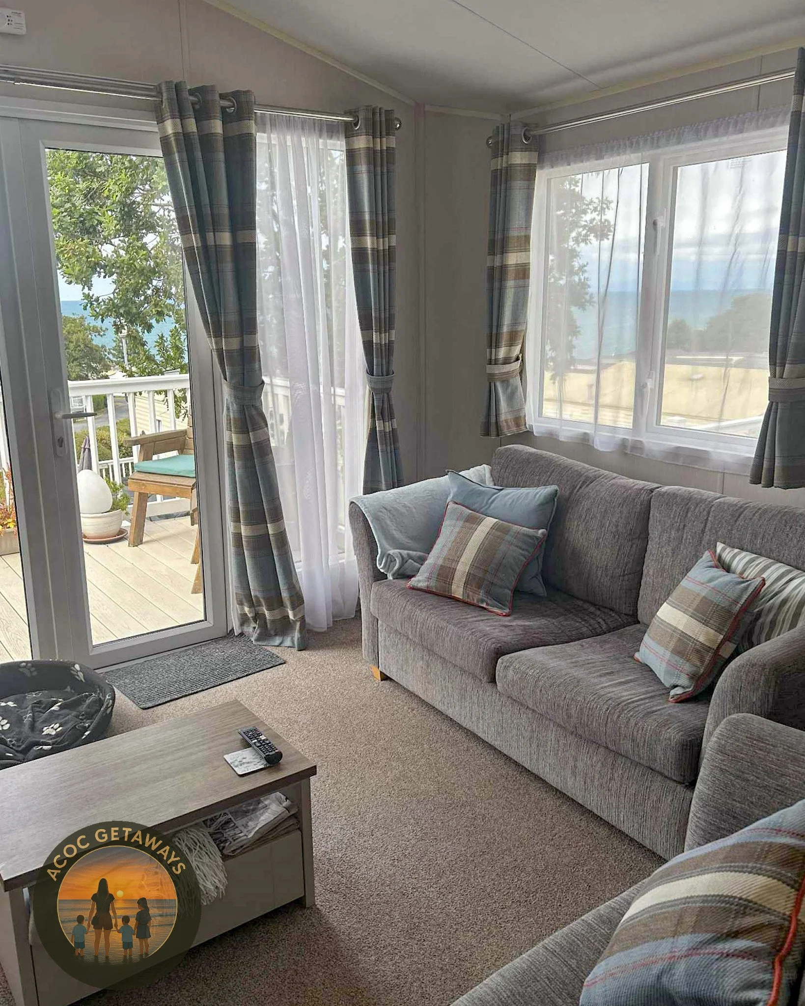 Living room with a gray sofa, plaid-patterned pillows, a sliding glass door with curtains leading to a balcony, and a window with curtains showing an outdoor landscape with trees and water.