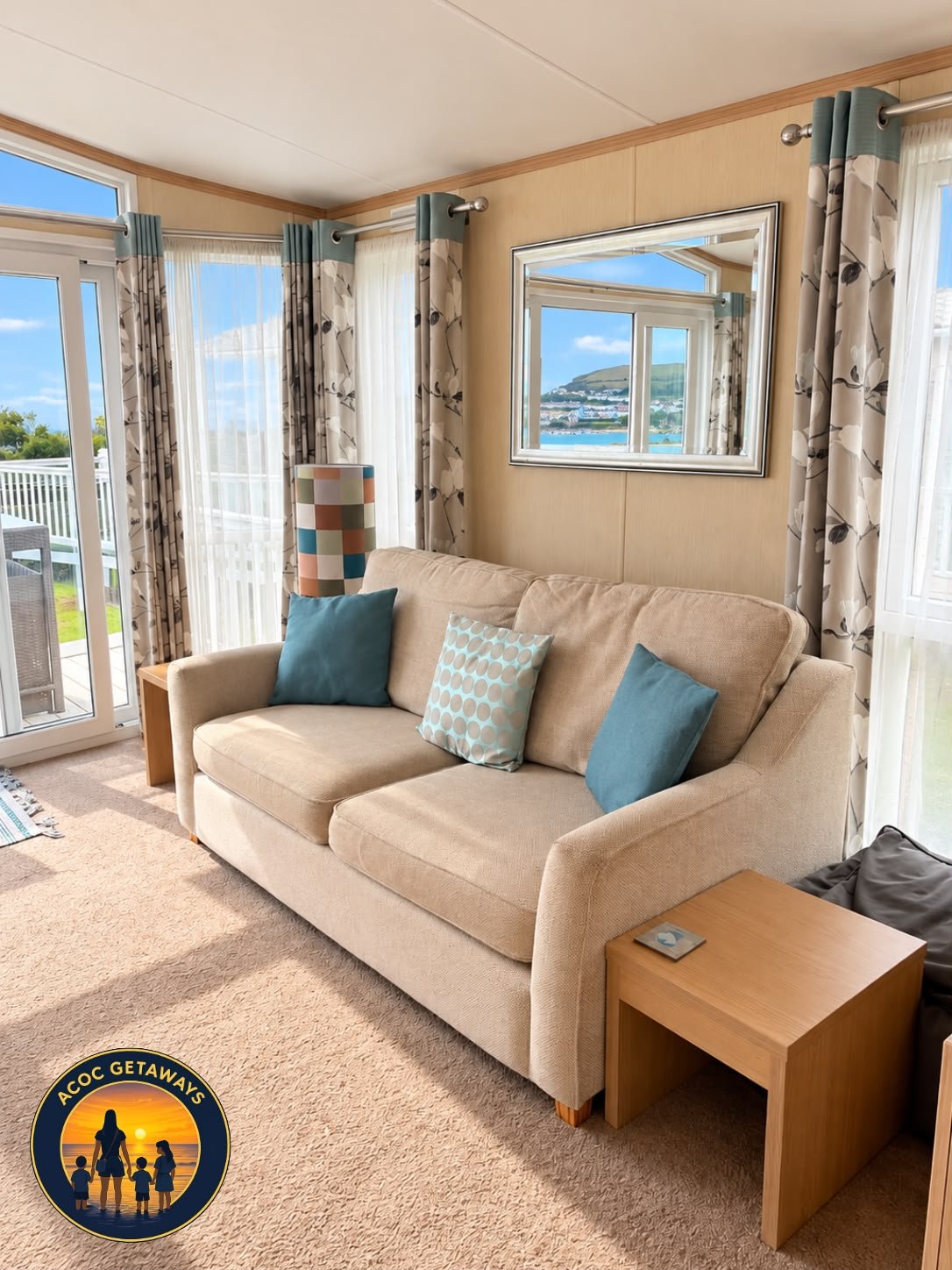 Living room with beige sofa, three decorative pillows, a lamp with a checkered shade, a framed mirror on the wall, and large windows with patterned curtains overlooking a balcony and a scenic landscape with water and hills.