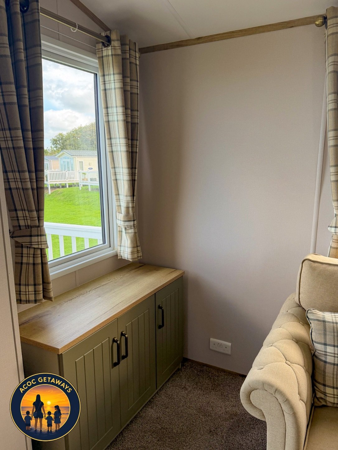 A small room with beige walls, a window with plaid curtains, a green cabinet with a wooden top, and a beige tufted armchair with plaid pillows.