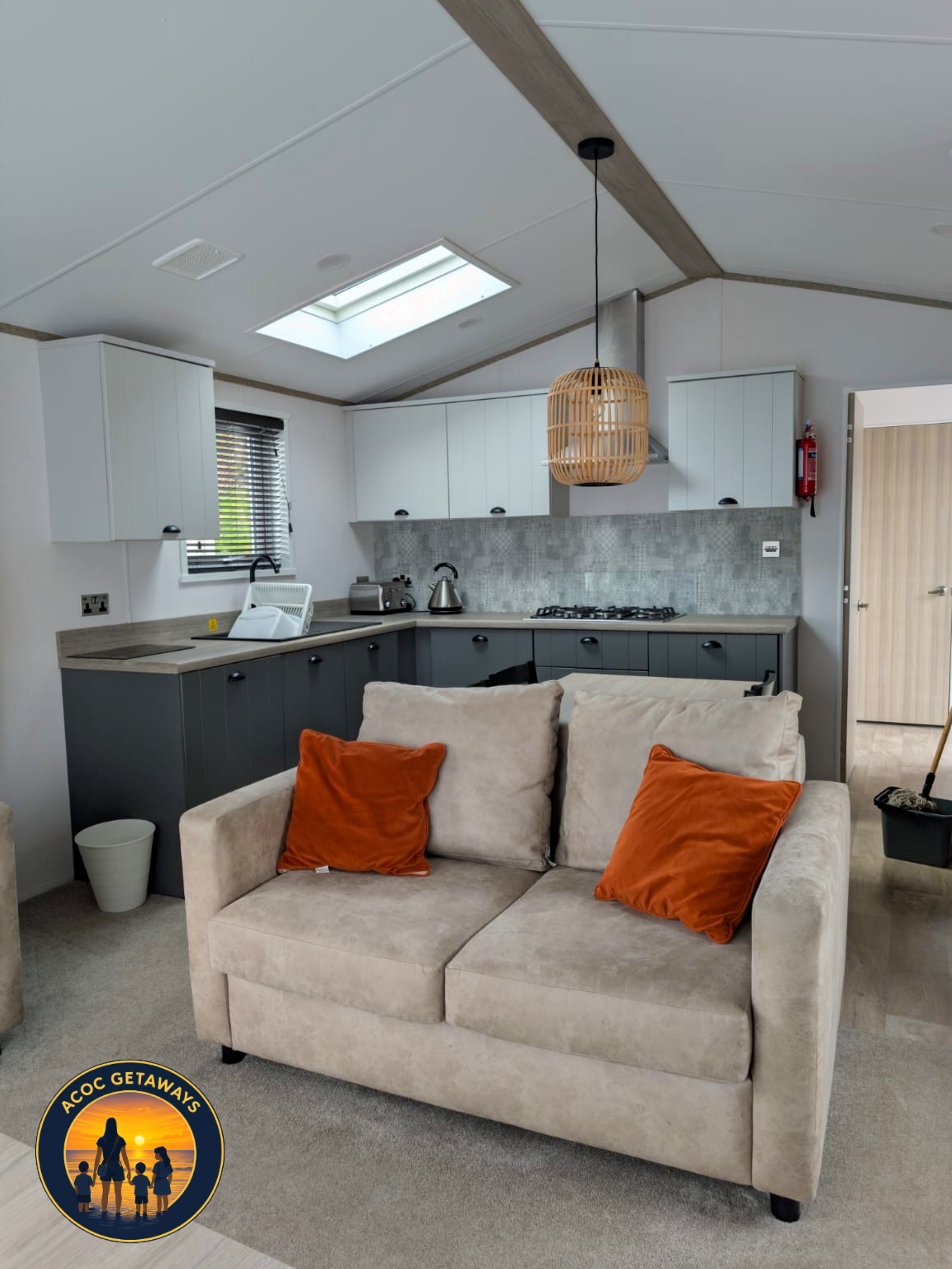A beige sofa with orange throw pillows in a living room with a kitchen in the background, a skylight, and a woven pendant light.