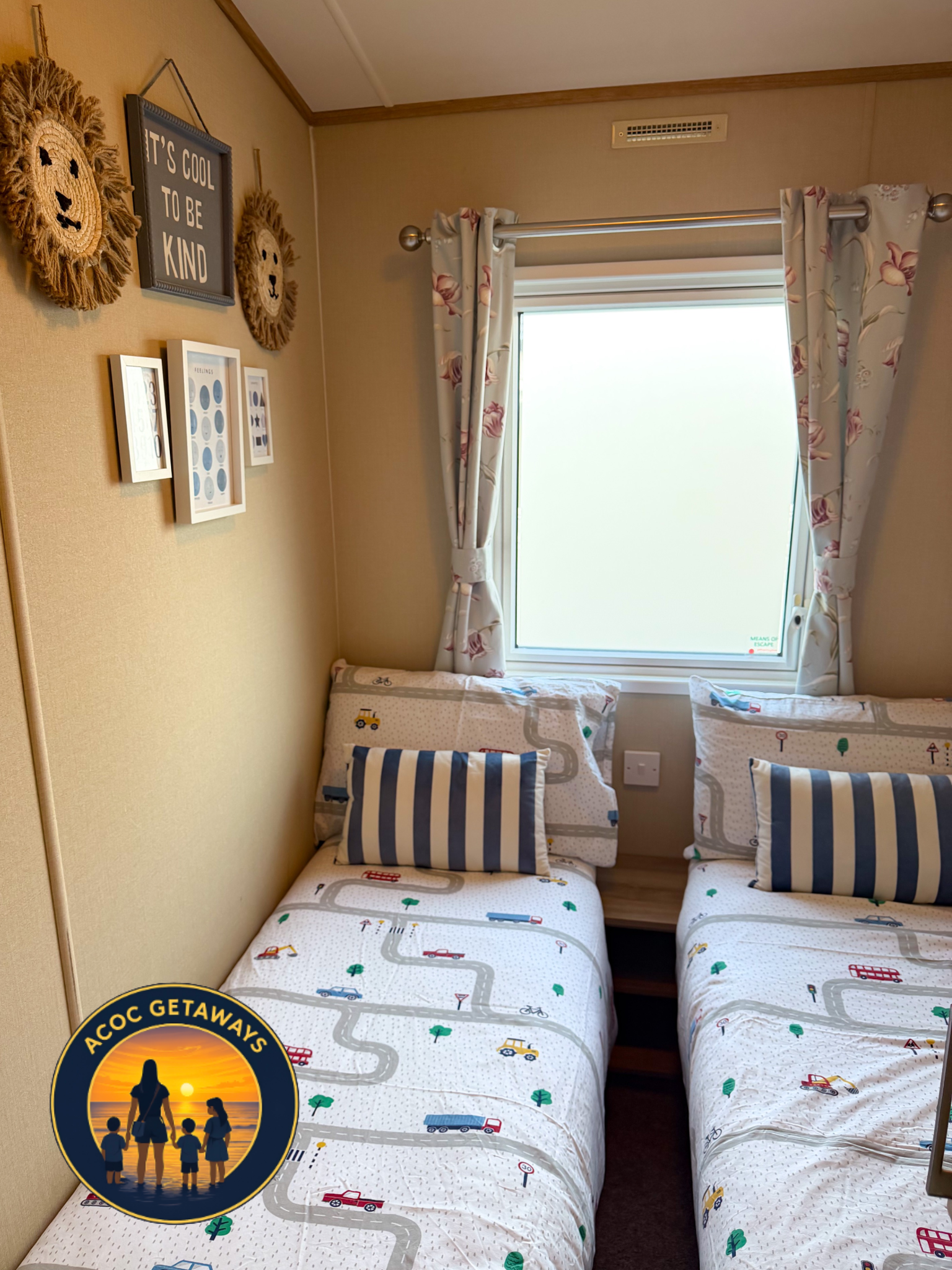 A cozy bedroom with two single beds, each with a striped blue and white pillow and bedding featuring a road and vehicle design. There is a window with light floral curtains, and wall decor including a framed sign reading 'It's cool to be kind,' paper