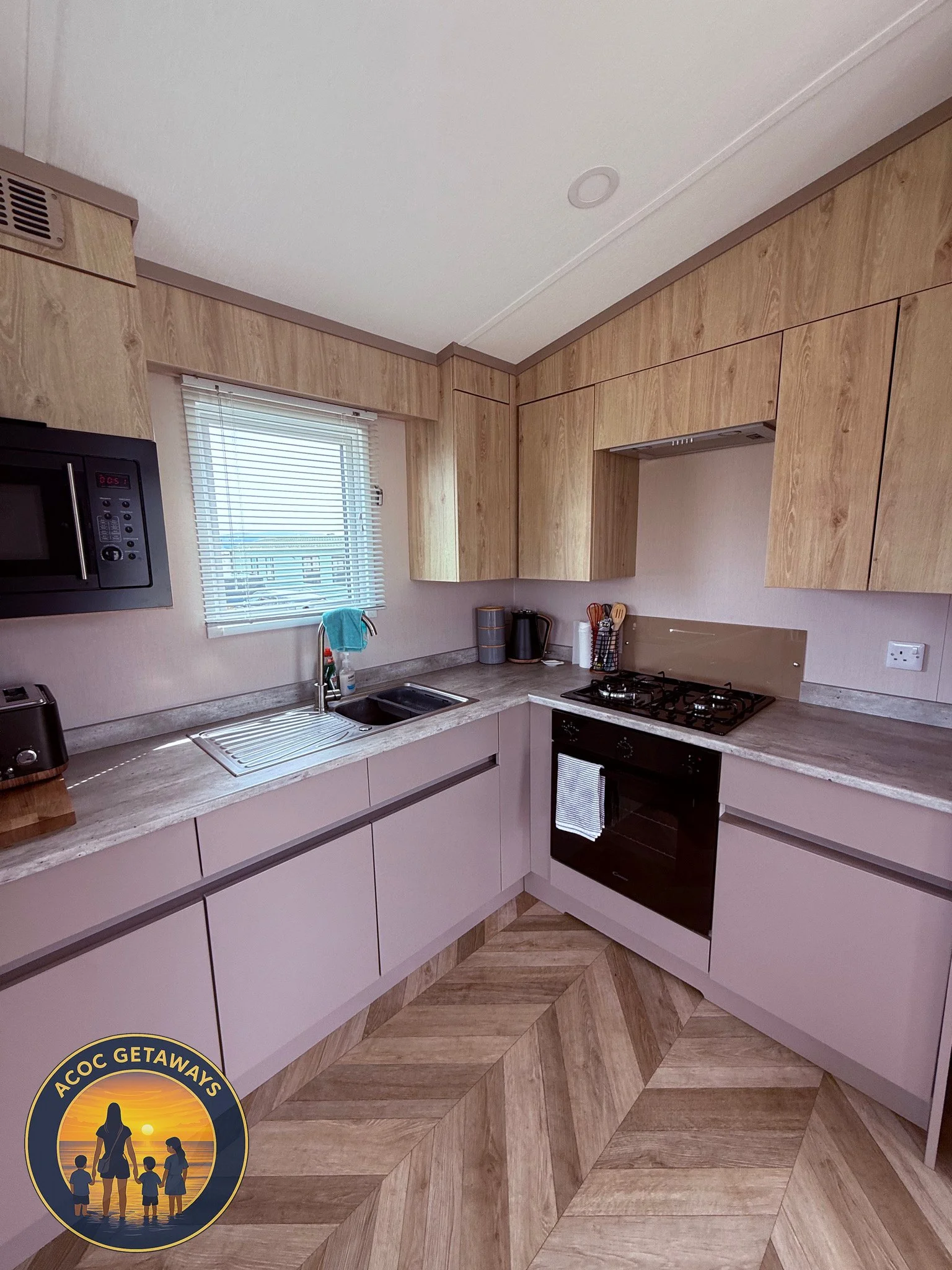 Modern kitchen with light wood cabinets, a small window with blinds above the sink, black microwave, toaster, teapot, utensils, and a gas stove with oven, featuring a wood and laminate pattern on the floor.