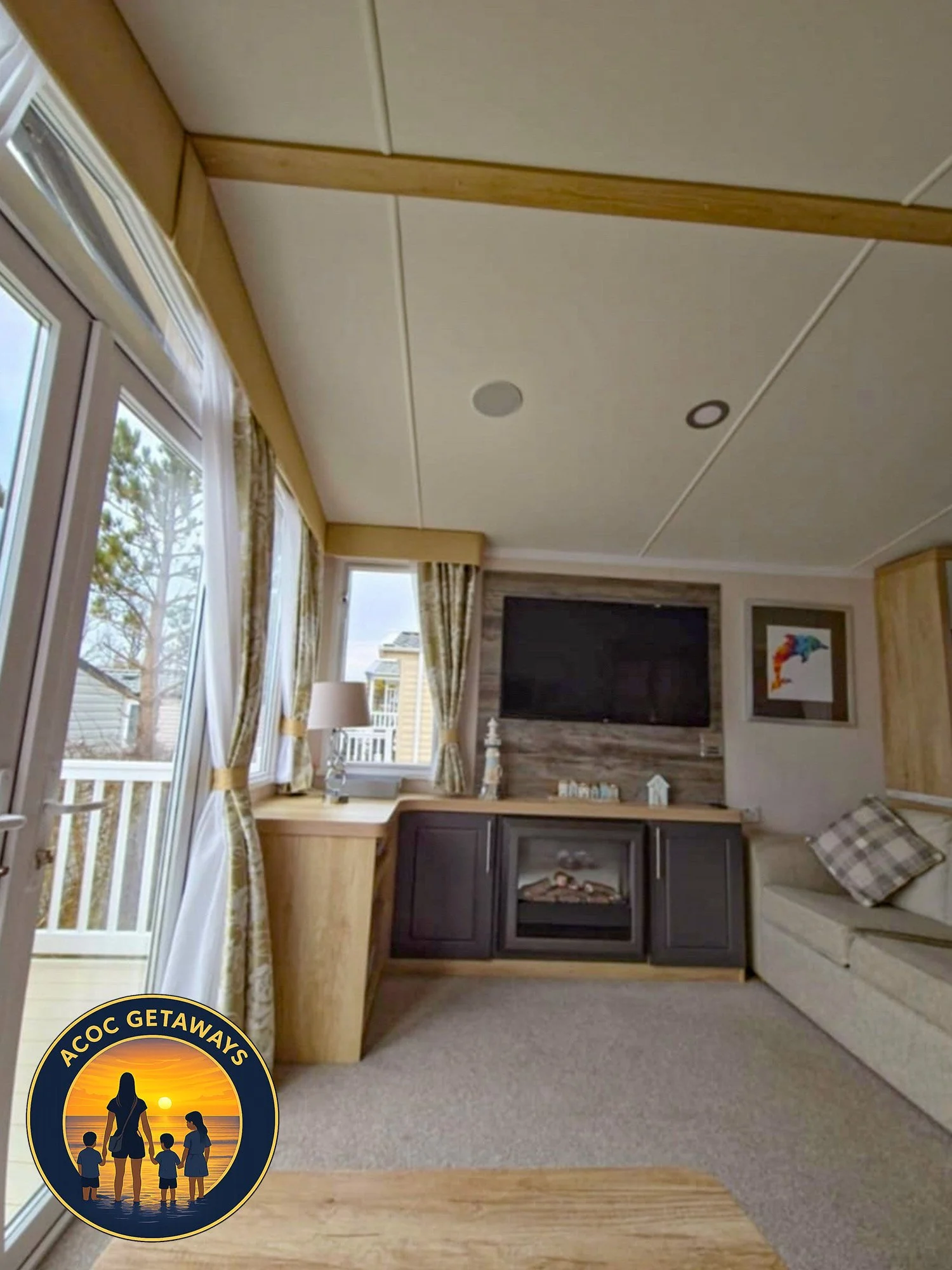 Interior of a living room with large windows, curtains, a mounted flat-screen TV, a sofa with checkered pillows, and wooden accents.