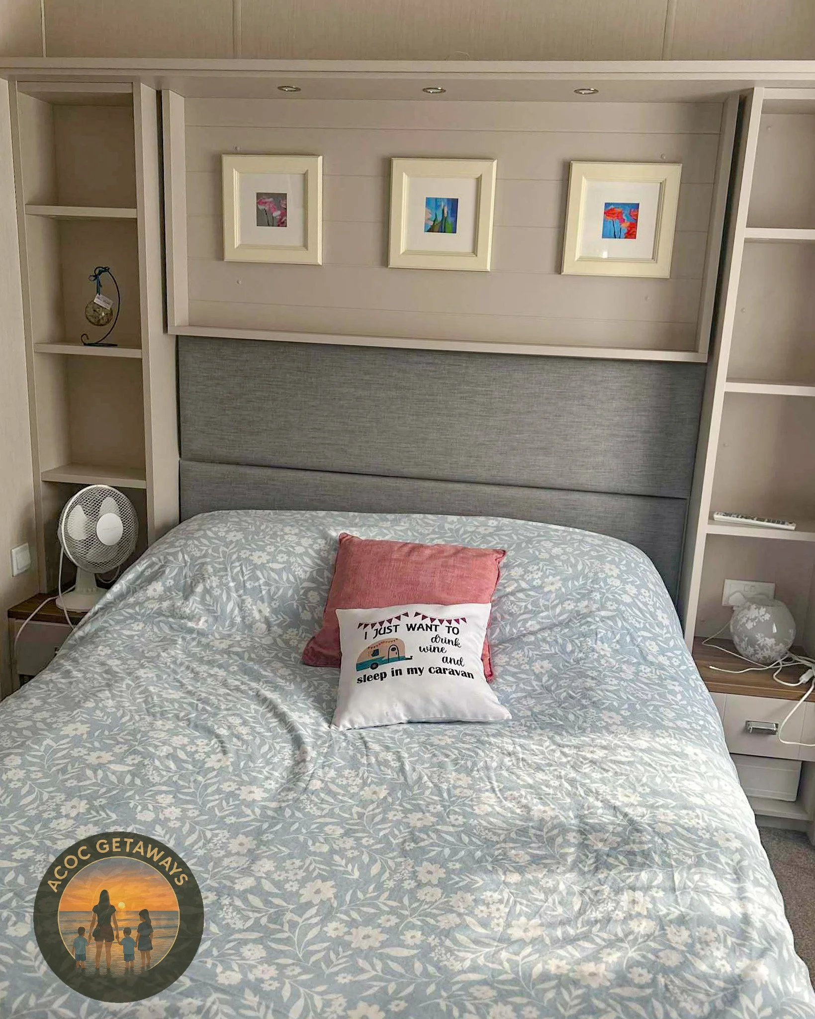 A bedroom with a bed that has a gray headboard, white floral patterned bedsheet, red pillow, and a white pillow with writing and small illustrations. There are three framed pictures above the bed and shelves on both sides. A small fan is on the left nightstand, and a lamp with a white base is on the right nightstand.