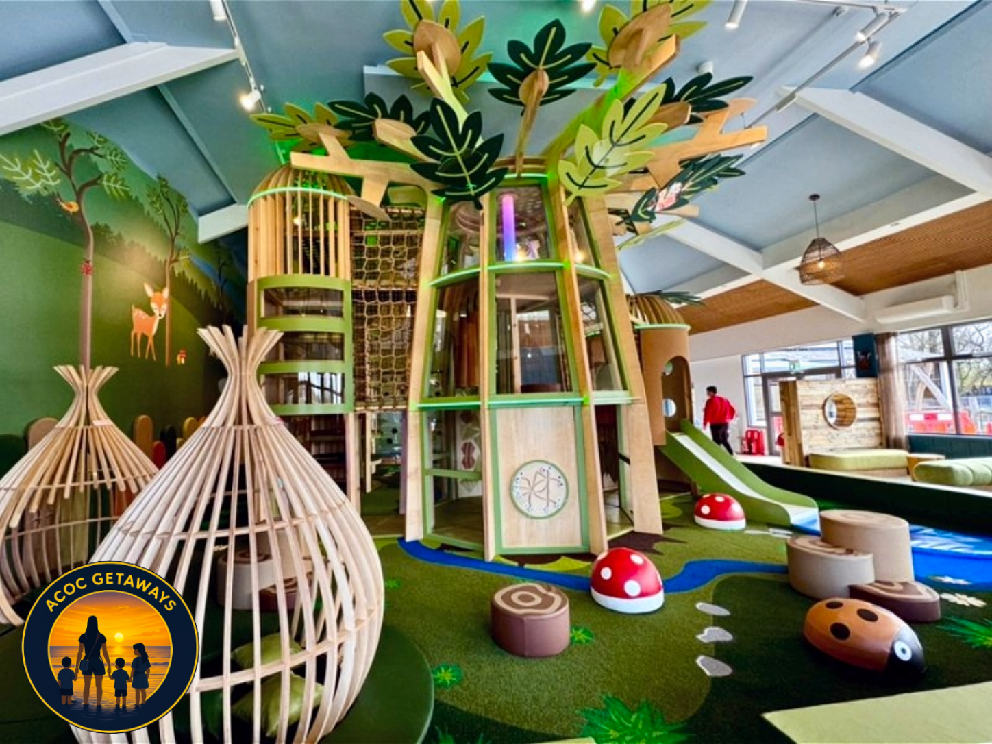 Indoor playground featuring a large wooden tree structure with a slide, climbing nets, and various play elements, surrounded by soft flooring with mushroom and log shapes, and decorative forest-themed wall art.