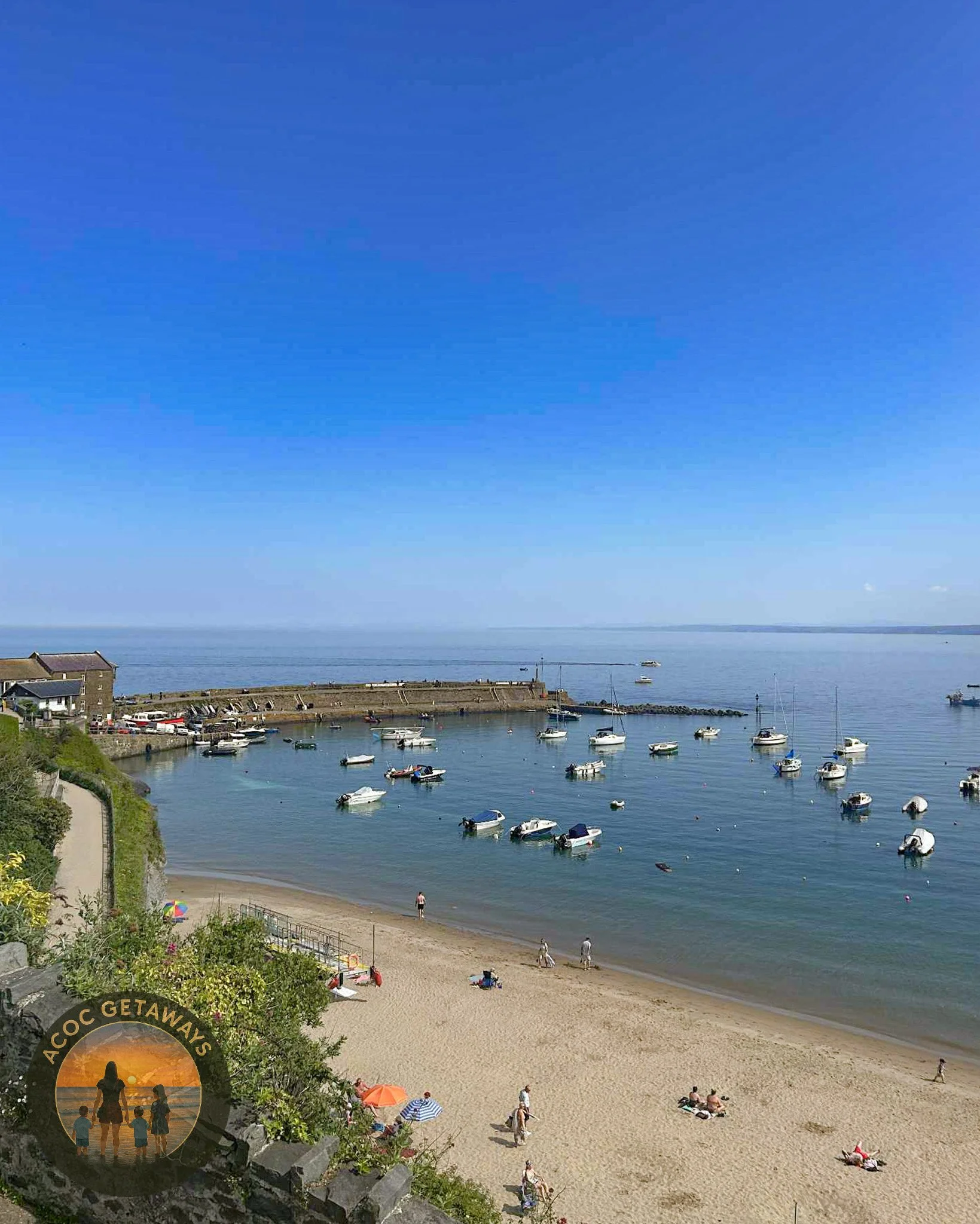 A peaceful beach scene with a sandy shoreline and boats anchored in calm water under a clear blue sky. People are walking and relaxing on the beach. A small harbor with buildings and greenery is visible on the left side of the image.