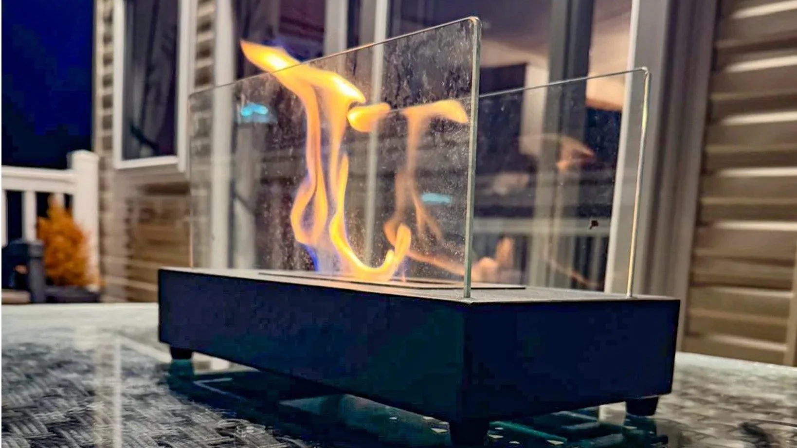 A tabletop patio heater with a glass enclosure and visible flame inside, on a granite table outdoors at night, with a wooden house and a window in the background.
