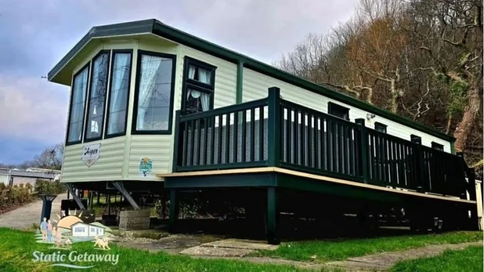 A small, elevated vacation home with white siding and black accents, featuring large windows and a wrap-around porch, situated on a grassy lot with trees in the background.